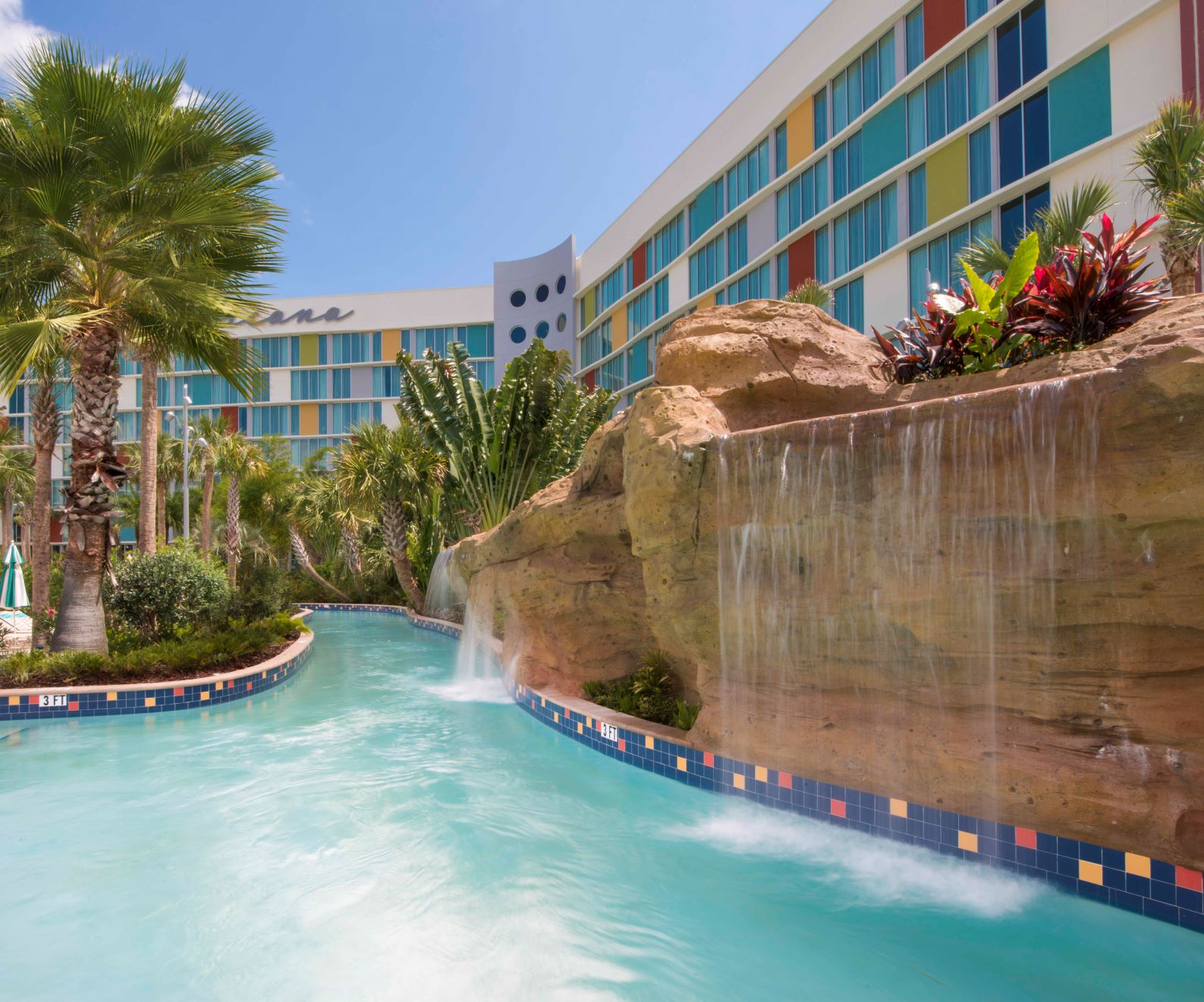 The lazy river at Universal Cabana Bay Beach Resort on a clear day. Turquoise water curves between a tiled edge and a tall artificial rock wall with water sheeting down it into the river. Palm trees and tropical planting line the left bank, and the hotel tower rises behind in white with panels of teal, yellow and red, with part of a large script sign reading Cabana visible on the far wing.