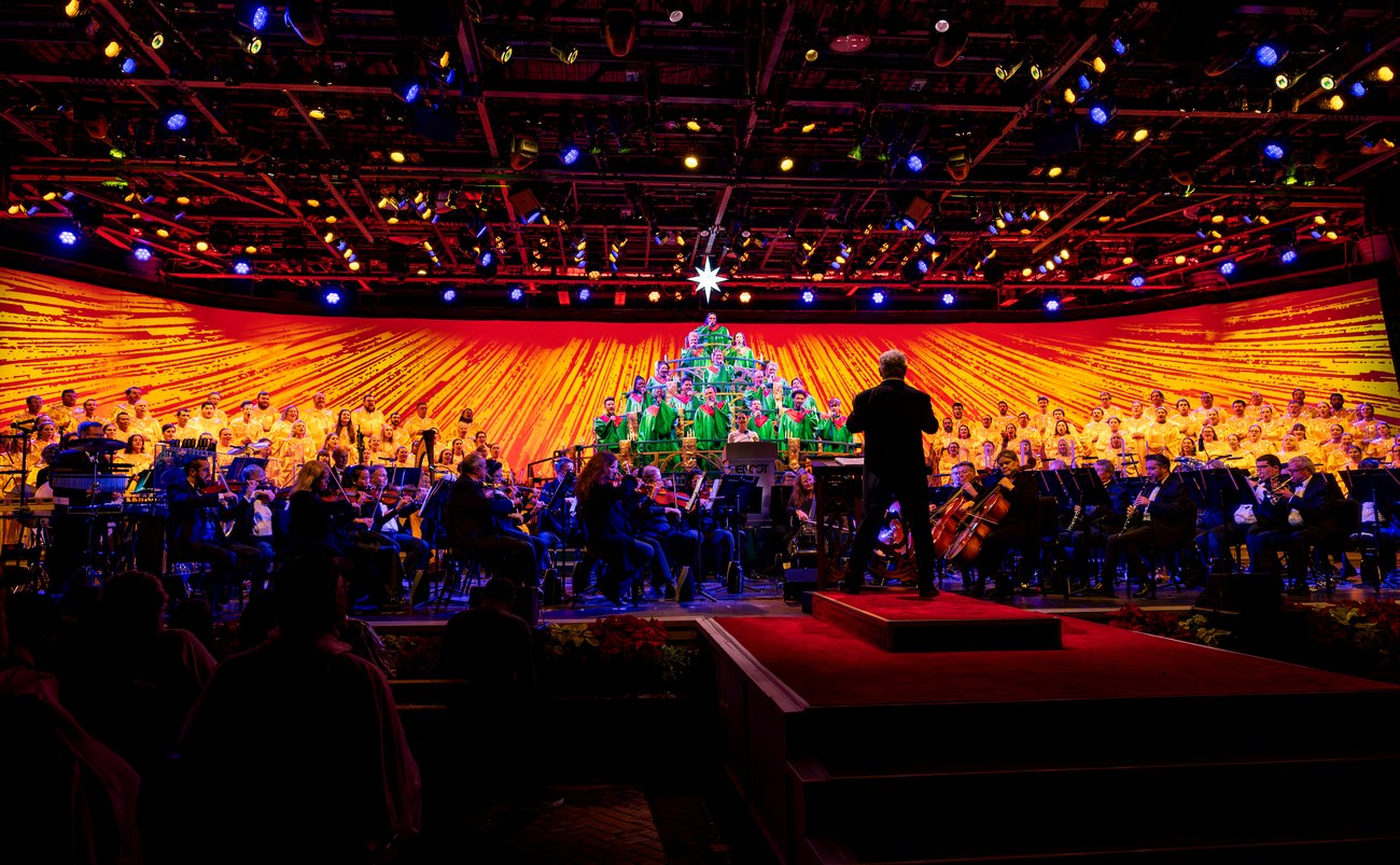 The Candlelight Processional at EPCOT's America Gardens Theatre, with a conductor on a riser in front of a full orchestra, a choir in yellow robes filling the stage, and singers in green robes arranged in a Christmas tree shape at center, backed by a red and gold sunburst backdrop topped with a star.