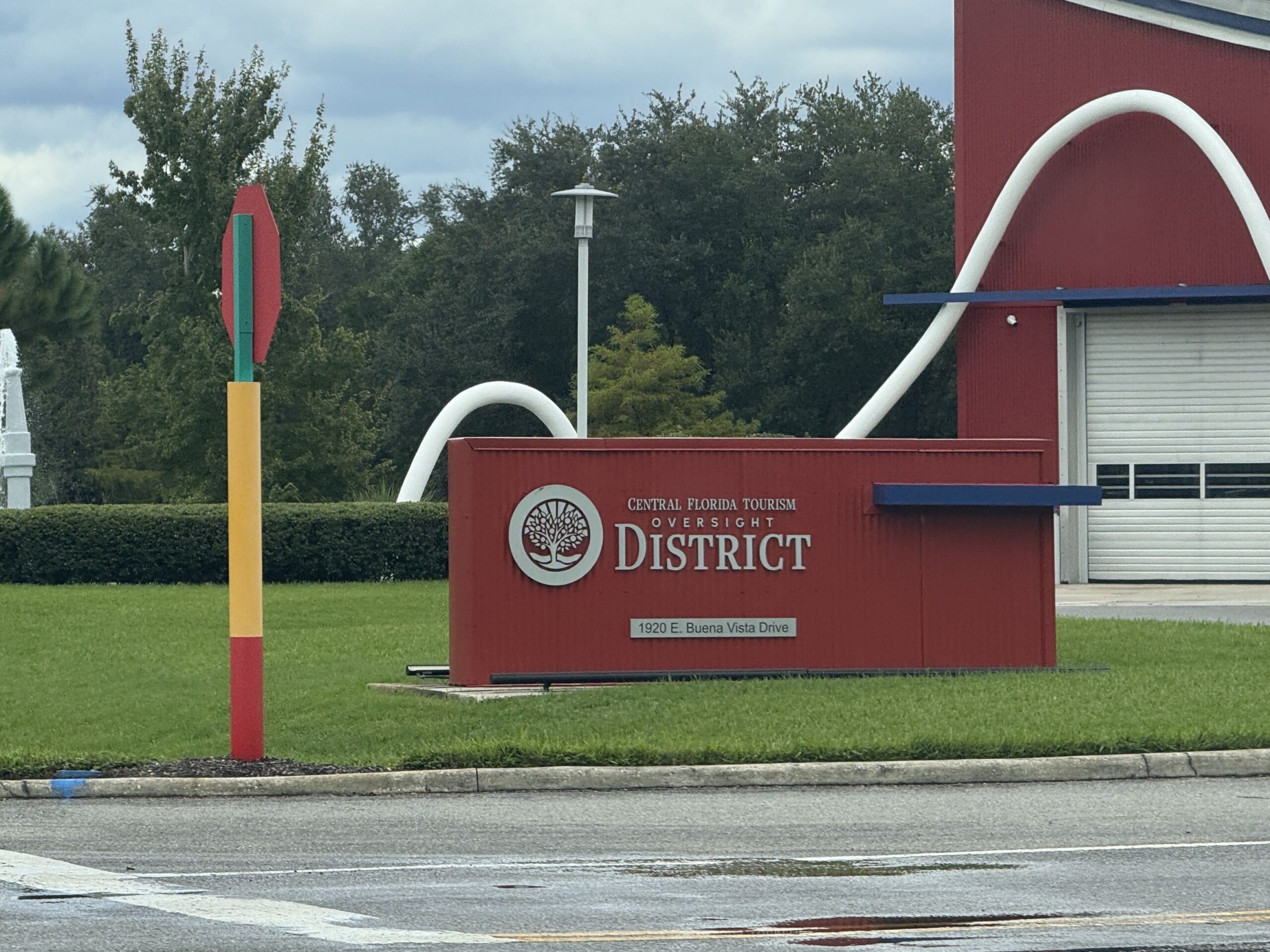 A low red monument sign on a lawn beside a wet road, in front of the red fire station wall and part of its white hose sculpture. The sign carries a circular silver logo of a tree and the words CENTRAL FLORIDA TOURISM OVERSIGHT DISTRICT in silver capitals, with a small plaque below reading 1920 E. Buena Vista Drive. A tall striped pole with a stop sign stands to the left.