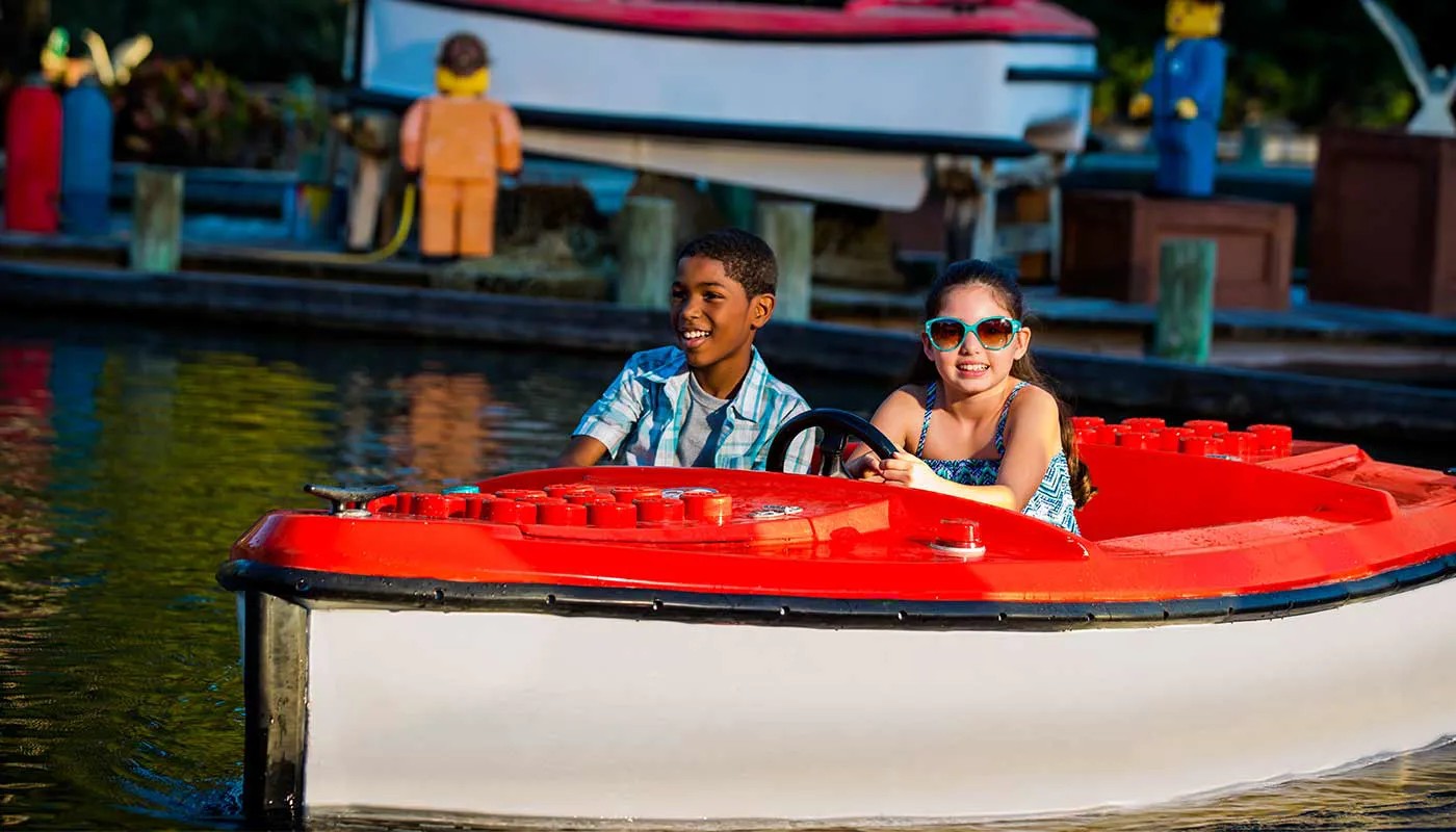 Two children ride a small motorized boat with a white hull and a bright red deck molded with oversized LEGO studs, steering along a green tinted waterway in warm low sunlight. A boy in a pale checked shirt sits at the left and a girl in turquoise sunglasses and a patterned top holds the black steering wheel. Behind them a wooden dock carries an orange LEGO minifigure model, a blue and yellow one, and a larger white and red boat model.