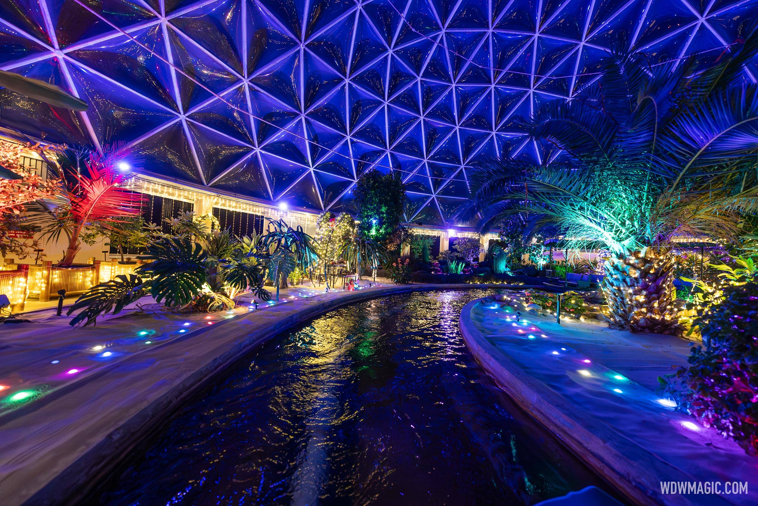 A greenhouse inside The Land at night, its glass dome lit deep blue and purple above the boat canal, with palms and planting beds edged by small colored path lights.