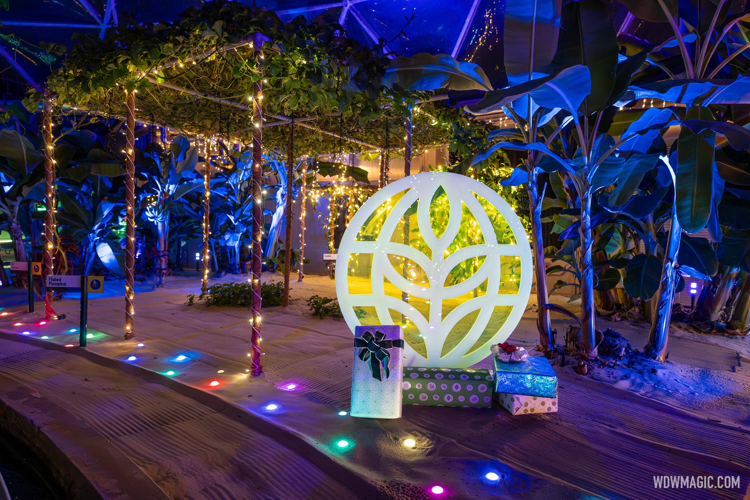 The illuminated white leaf logo sculpture of The Land Pavilion glowing on a sand bed at night, with wrapped gift boxes stacked in front of it and banana plants lit blue behind.