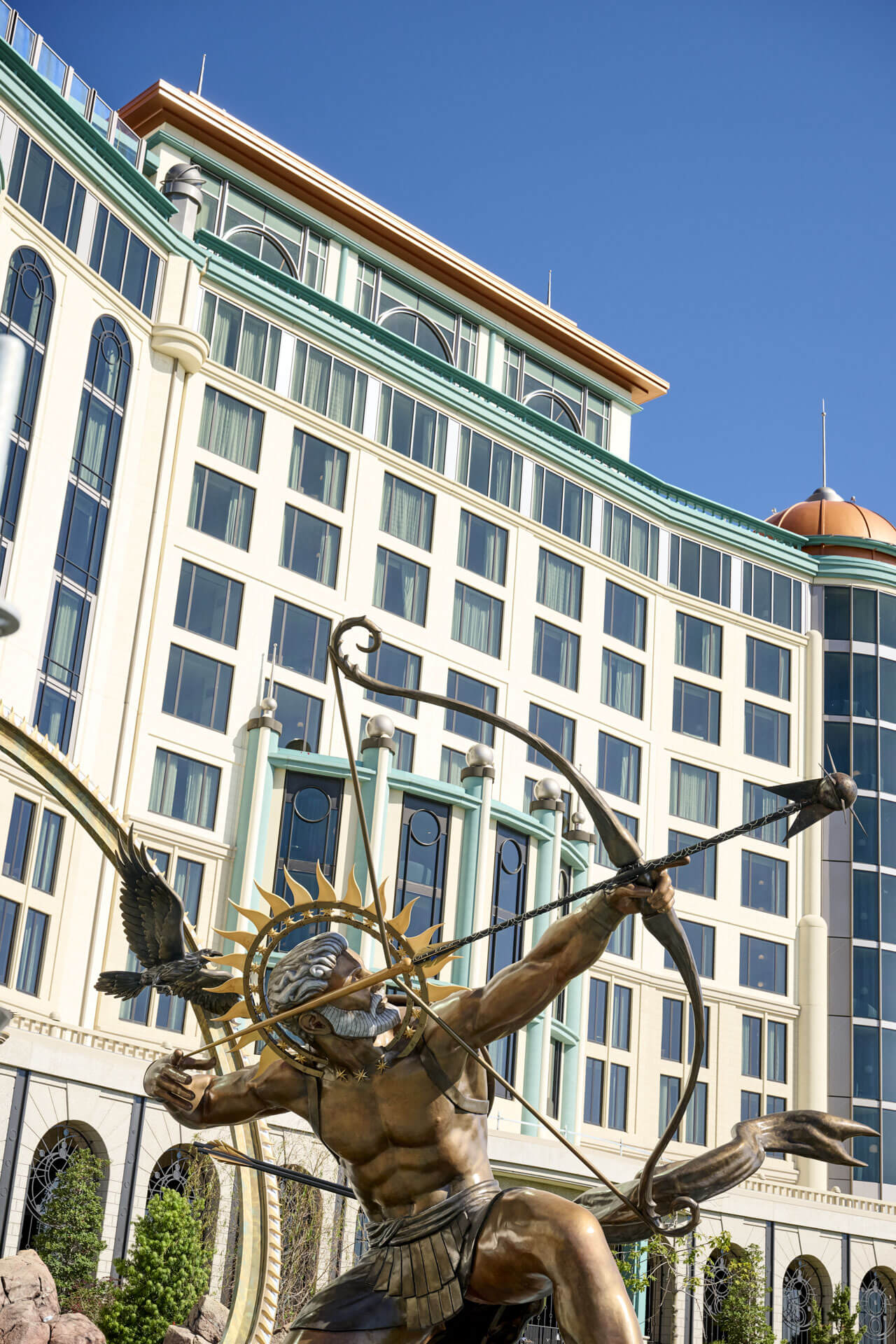 A bronze statue of an archer with a golden sunburst halo behind his head drawing a bow, with a bronze bird in flight beside him, photographed from below against a cloudless blue sky. Behind rises the cream and pale green facade of Universal Helios Grand Hotel, with tall arched and rectangular windows, a green cornice, a copper colored dome at the right, and mint green columns topped with white globe lamps around its entrance.