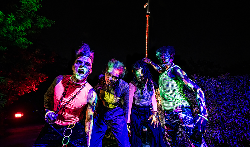Four Howl-O-Scream scare actors posed at night in ultraviolet lighting, their skin and clothing painted in glowing pink, green, yellow and blue. One in a pink tank top has a chain looped across the chest and is shouting at the camera, one wears a yellow crop top and blue trousers with mesh sleeves, and one at right in a green mesh vest leans on a tall pole. Dark foliage and a red light sit behind them.
