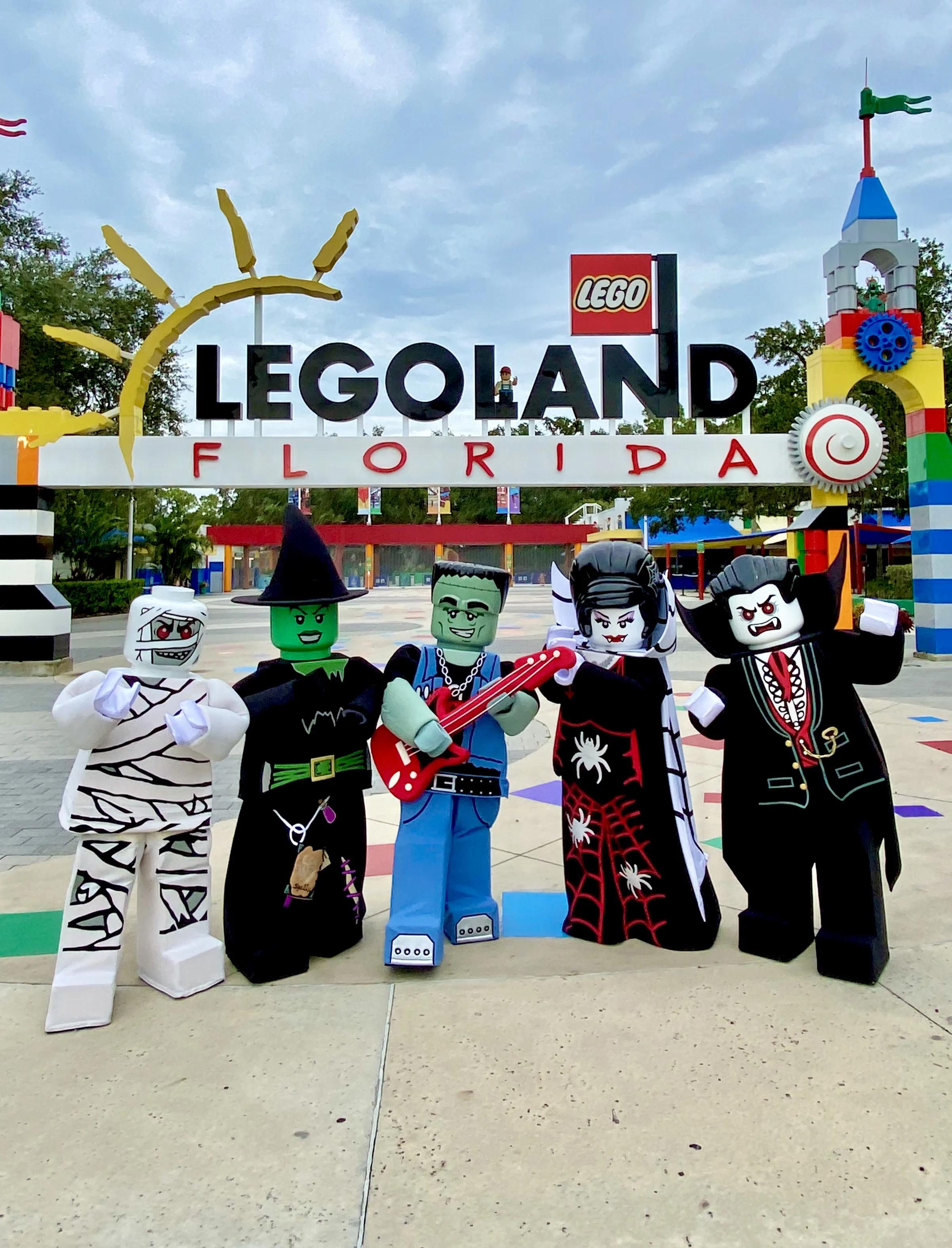 Five costumed LEGO character mascots, a mummy, a green faced witch in a pointed hat, a green monster rocker holding a red toy guitar, a fanged character in a black gown covered in red webs and white spiders, and a vampire in a black cape, posed together in front of the LEGOLAND FLORIDA entrance sign under a cloudy sky.