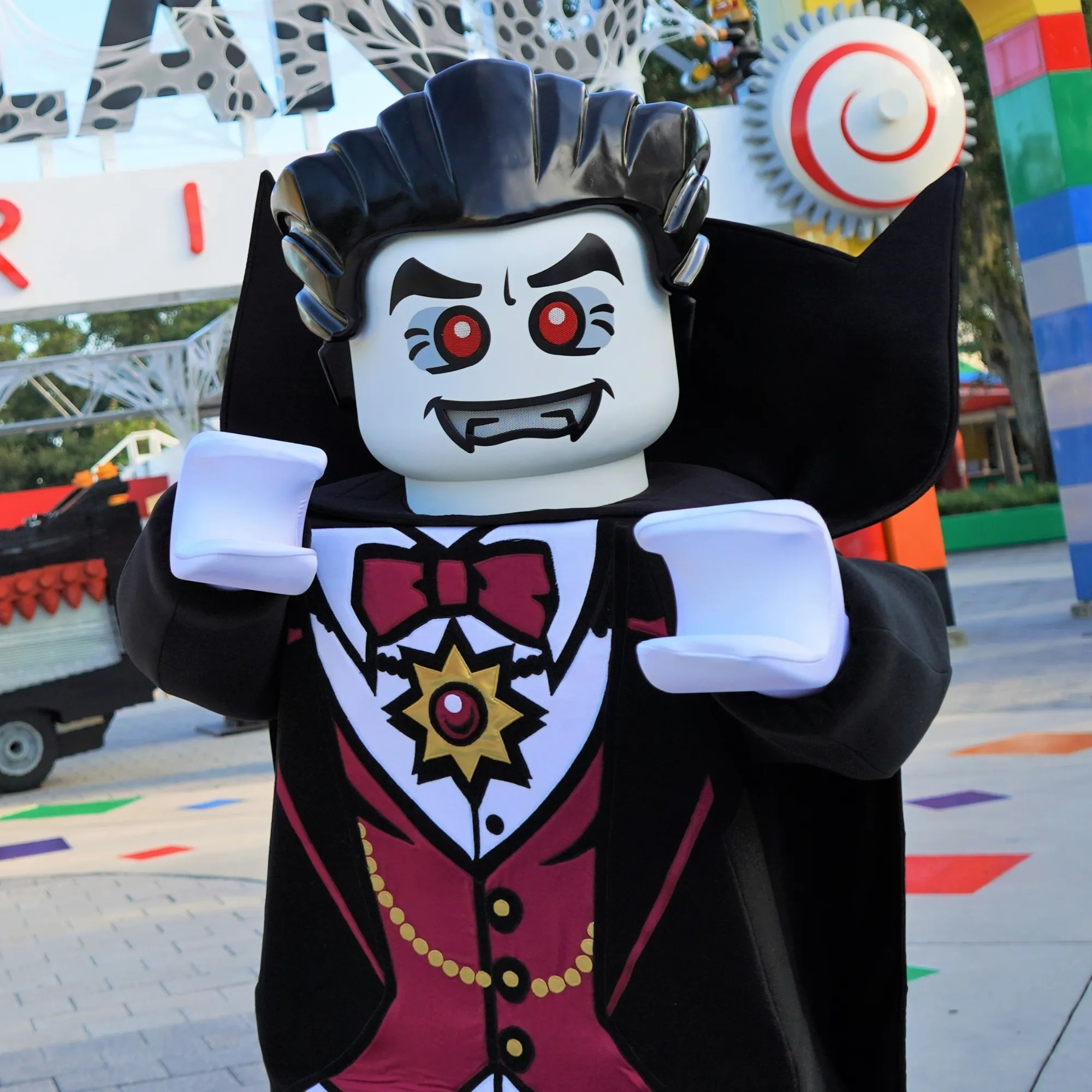The LEGO Lord Vampyre walkaround character outdoors in daylight, arms raised. He has a glossy molded black pompadour, a white face with red eyes and fangs, a black cape with a high pointed collar, a dark red waistcoat with gold buttons and chain, a maroon bow tie and a gold starburst medallion. Behind him are the letters of the LEGOLAND Florida entrance sign, a white and red spiral sculpture on a gear, and a rainbow striped column.