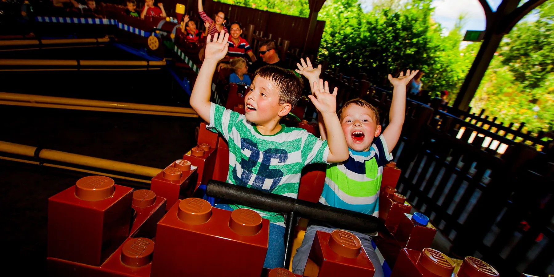 Two boys ride at the front of Merlin's Challenge with both arms thrown in the air and wide open mouths, one in a green and white striped shirt with a number on it and one in a blue and green striped shirt. Their car is built from oversized dark red LEGO bricks with visible studs and a black lap bar. More riders including adults sit in cars stretching back along the track behind them, beside a dark wooden fence and bright green foliage lit by sun.
