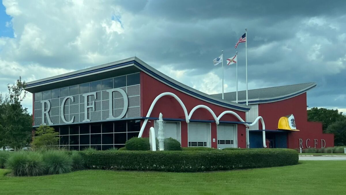 The same fire station photographed around 2023 under a grey and white cloudy sky. Large silver letters on the louvred panel spell RCFD, a smaller set of RCFD letters sits on the red wall at the far right end, and the yellow fire hat sculpture above them carries a pale shield with a dark crest on it. Three flags fly from poles behind the building and a clipped hedge and lawn run across the foreground.