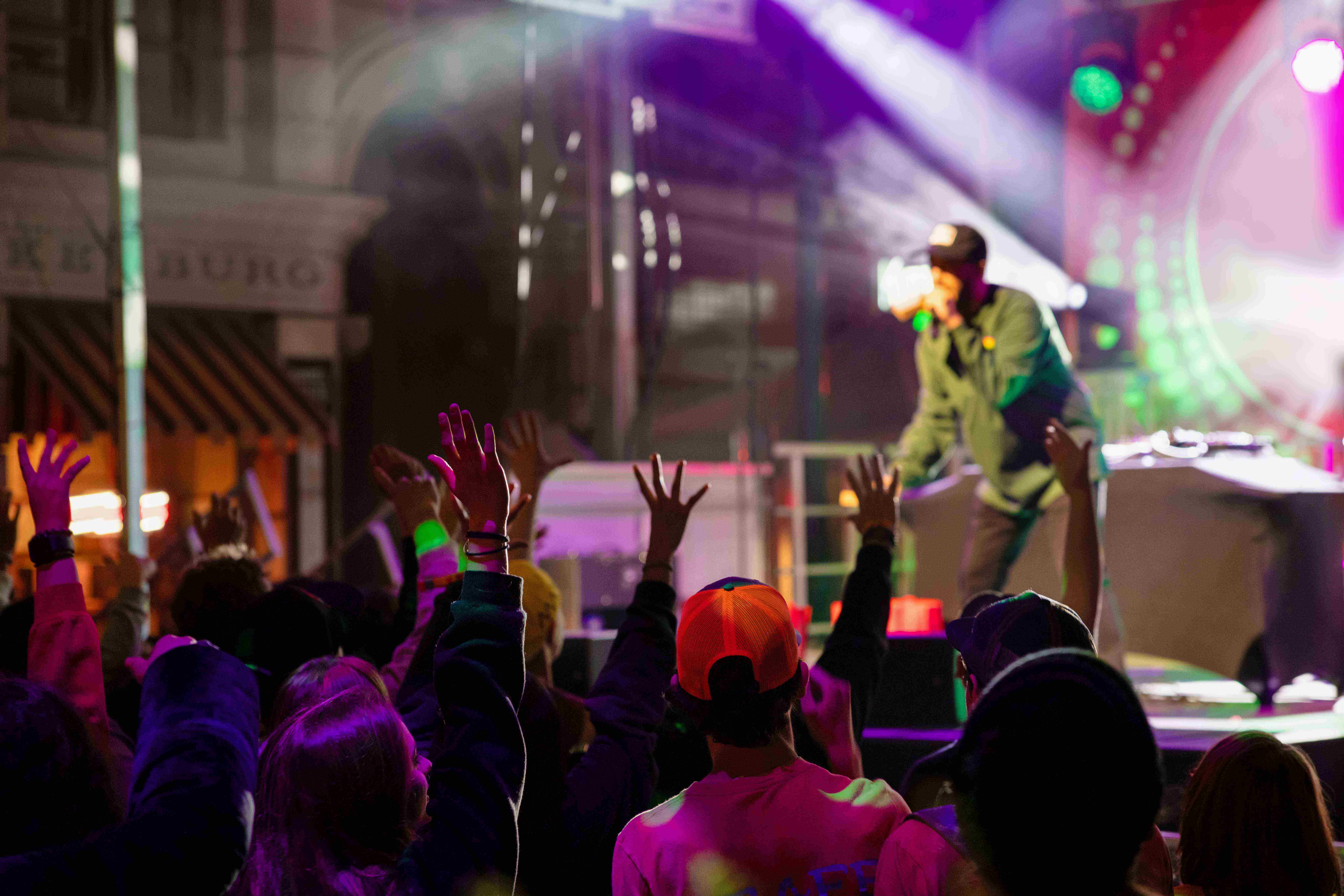 A night time Rock the Universe concert on an outdoor stage set up on a street of building facades at Universal Studios Florida, a performer in a light green jacket and dark cap singing into a handheld microphone under magenta and green stage lighting, photographed over a crowd with hands raised.