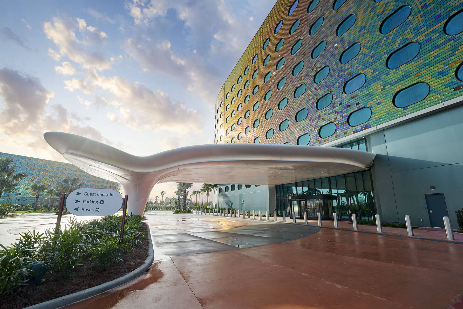 The guest entrance of Universal Terra Luna Resort at twilight, the pavement still wet. A sweeping white sculptural canopy on a single tapered column shelters the drive in front of a curved building clad in iridescent gold, green and blue tile with rows of large rounded oval windows. A directional sign at the left reads Guest Check-In, Parking and Buses, and a sister building in the same iridescent tile, in cooler blue and teal tones, is visible in the distance.