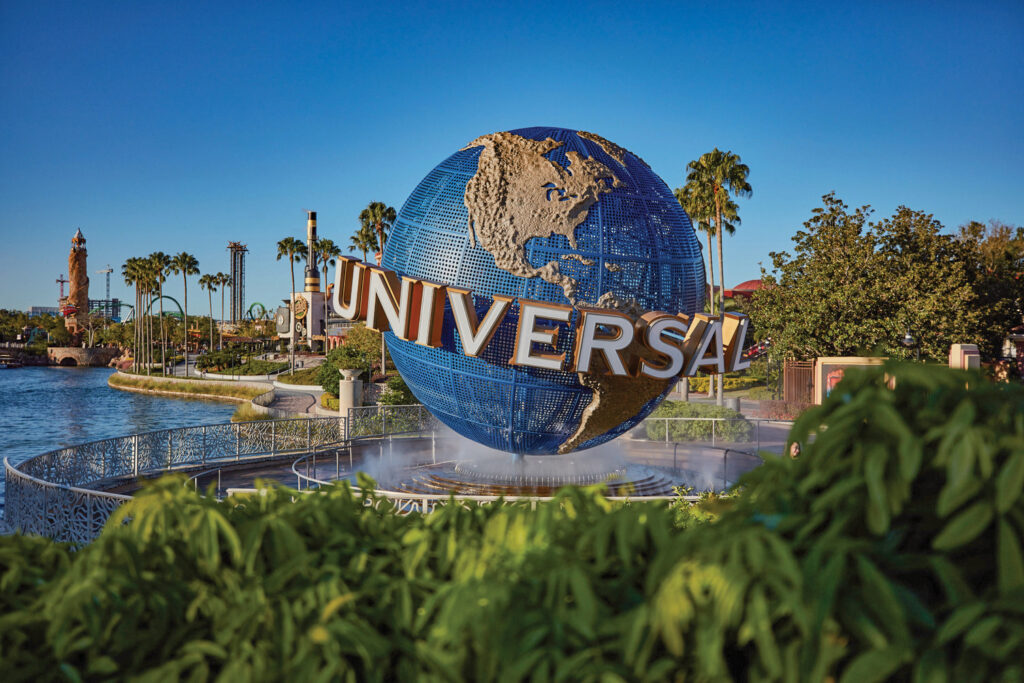 The rotating Universal globe at the resort entrance on a clear day, its blue latticework and raised gold and silver continents catching the sun, with UNIVERSAL in chrome letters across the middle. Water jets ring the base, palm trees and a lagoon sit behind it, and green foliage fills the foreground.