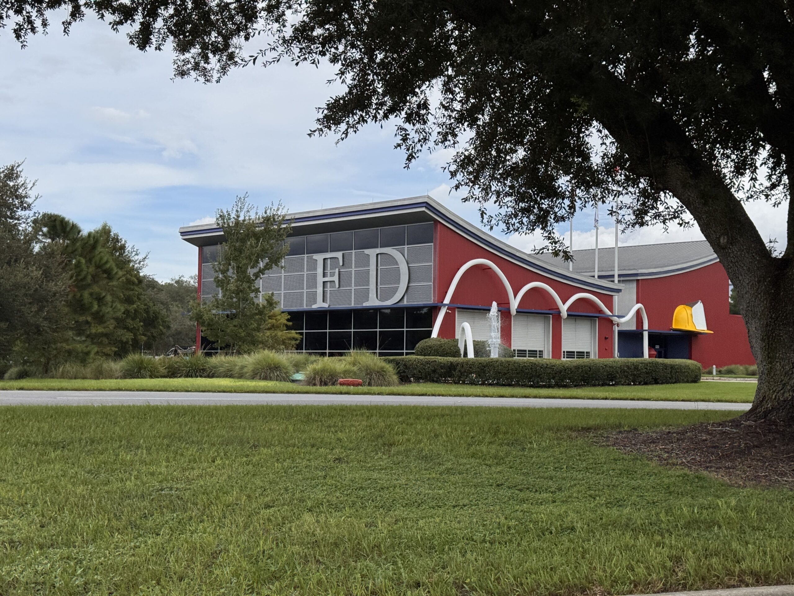 The Walt Disney World fire station photographed from across the road in daylight. The long red building has a swooping roofline with a blue fascia, a glass and grey louvred upper section carrying two large silver letters reading FD, a white hose sculpture looping three times over the engine bay doors, a small water fountain in front, and a large yellow fire hat sculpture mounted on the wall at the right end with a plain white shield on it. A live oak frames the right of the frame.