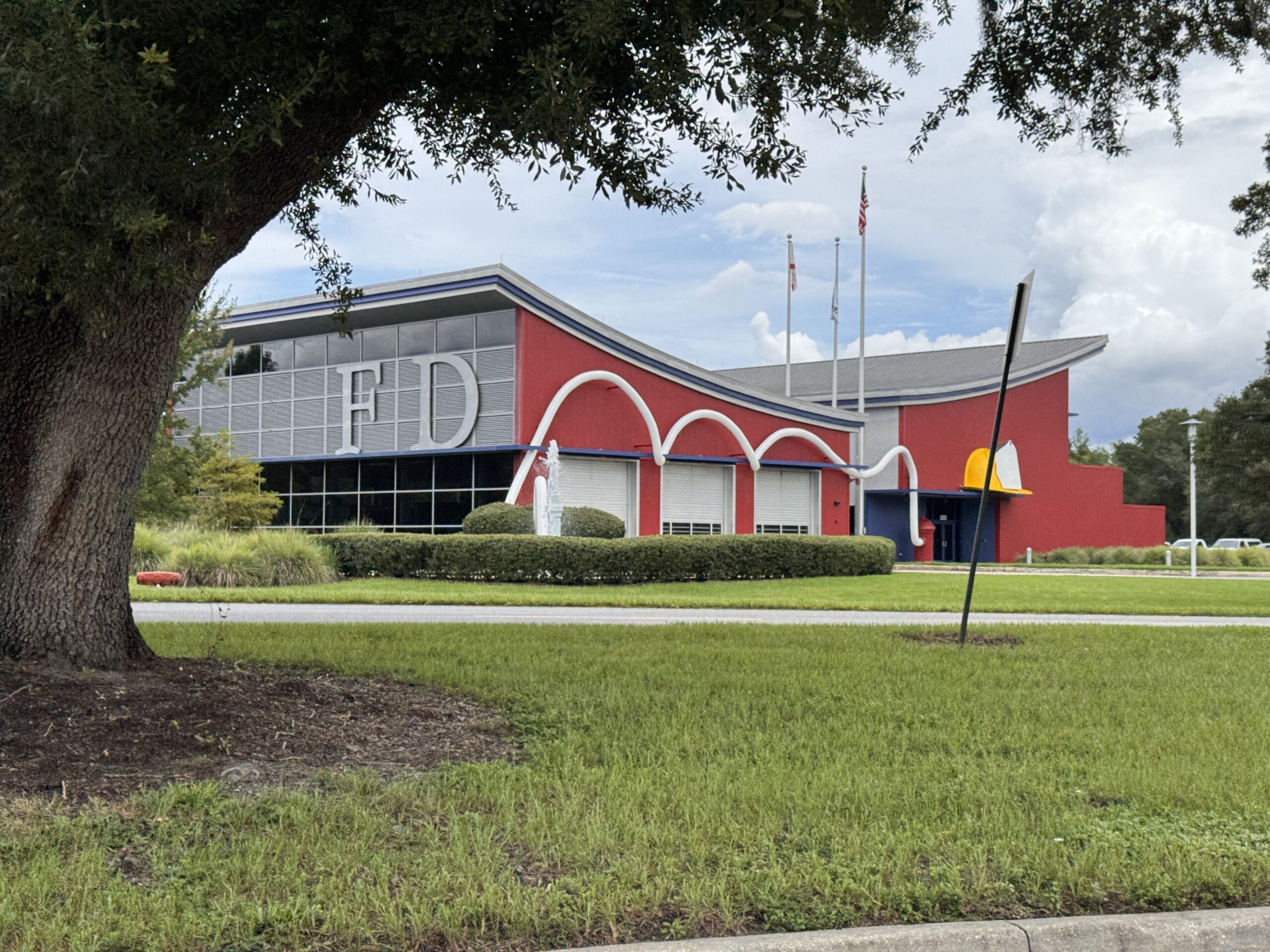 A wider view of the fire station from across the road, framed by the trunk and canopy of a large live oak on the left. The FD letters are visible on the louvred panel, the three white hose loops arc over three closed engine bay doors, the yellow fire hat with its white shield sits at the right end of the red building, and three flagpoles rise behind the roof, one flying the United States flag and one the Florida state flag.