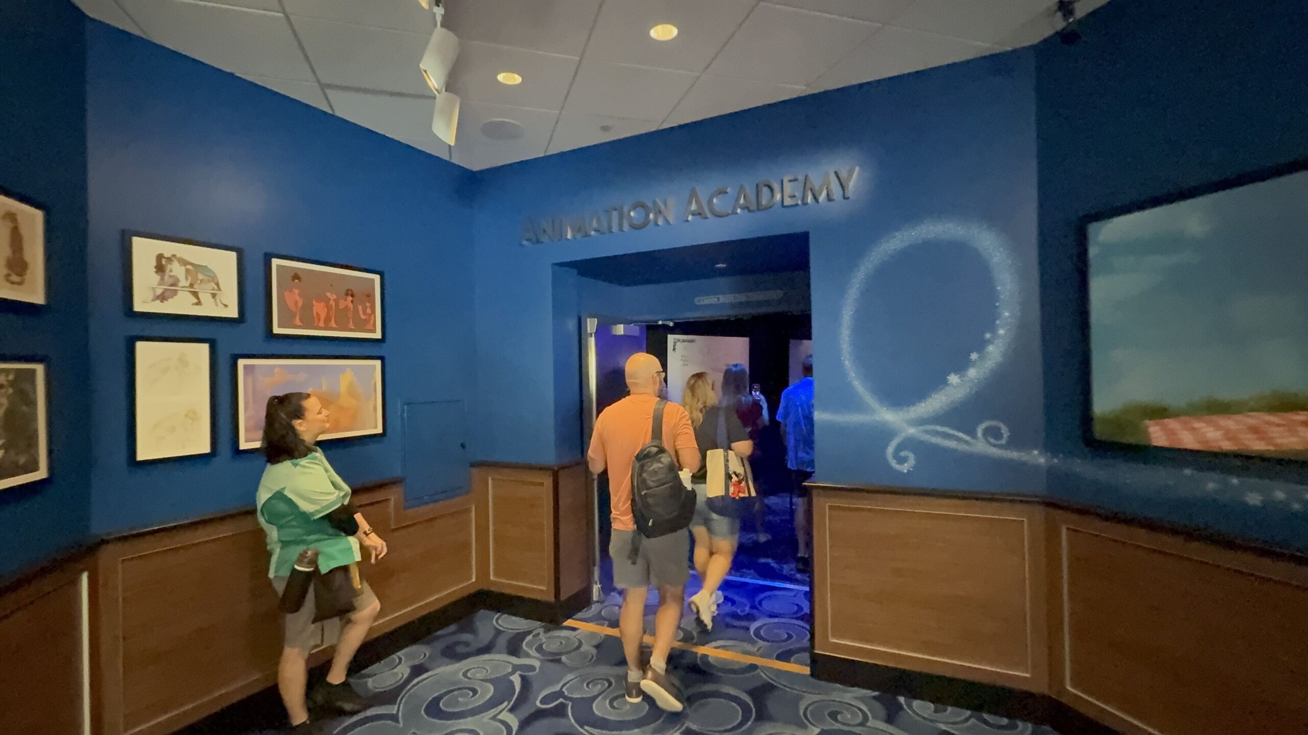 Guests walk through a blue gallery room hung with framed Disney artwork toward a doorway lettered ANIMATION ACADEMY, with a trail of snowy sparkles curling across the wall from a framed Frozen scene on the right