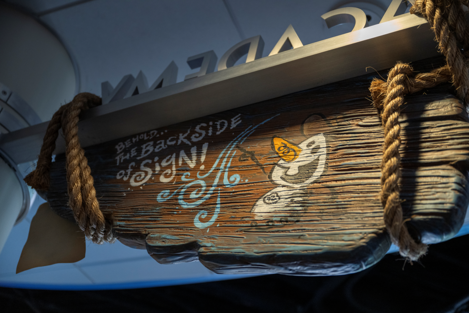 The reverse of a wooden hanging marquee slung on rope, hand lettered BEHOLD... THE BACKSIDE of SIGN! beside a sketched Olaf, with the metal letters of the ACADEMY sign visible behind it