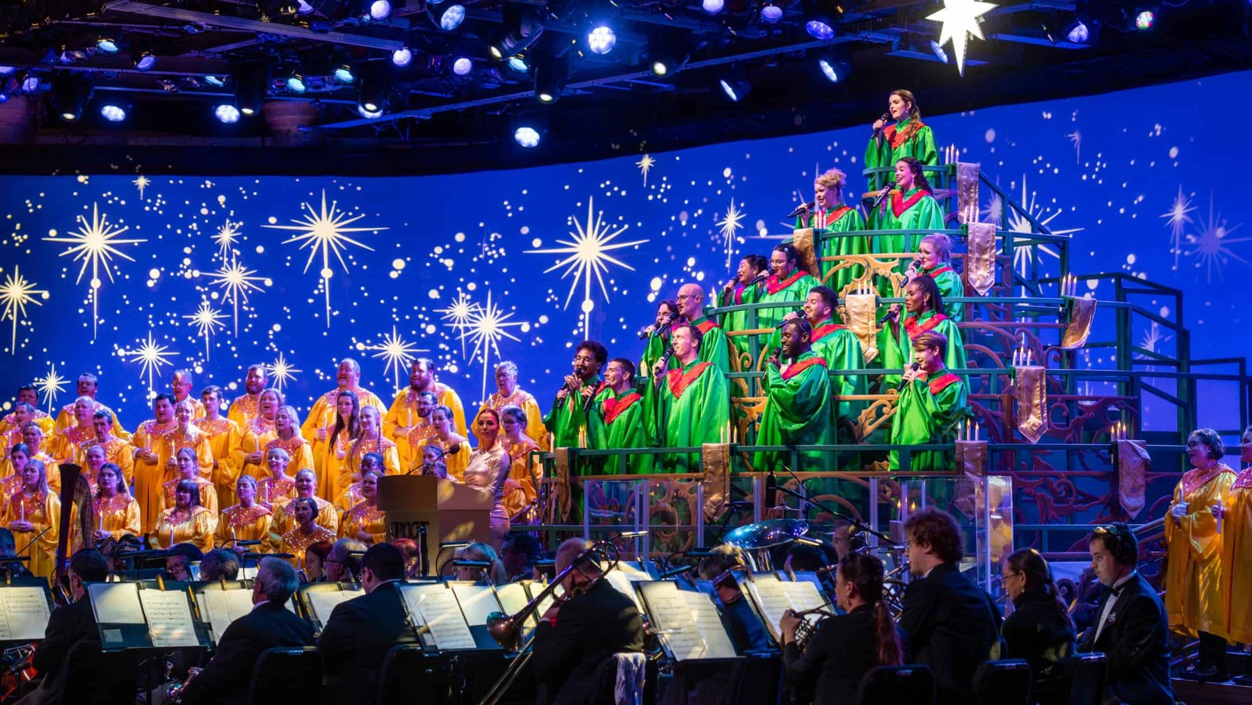 A closer view of the Candlelight Processional choir, with singers in green robes filling the tiered riser at right, choir members in gold robes holding lit candles at left, a woman in a gold gown at an EPCOT lectern, an illuminated star above, and the orchestra with sheet music in the foreground.