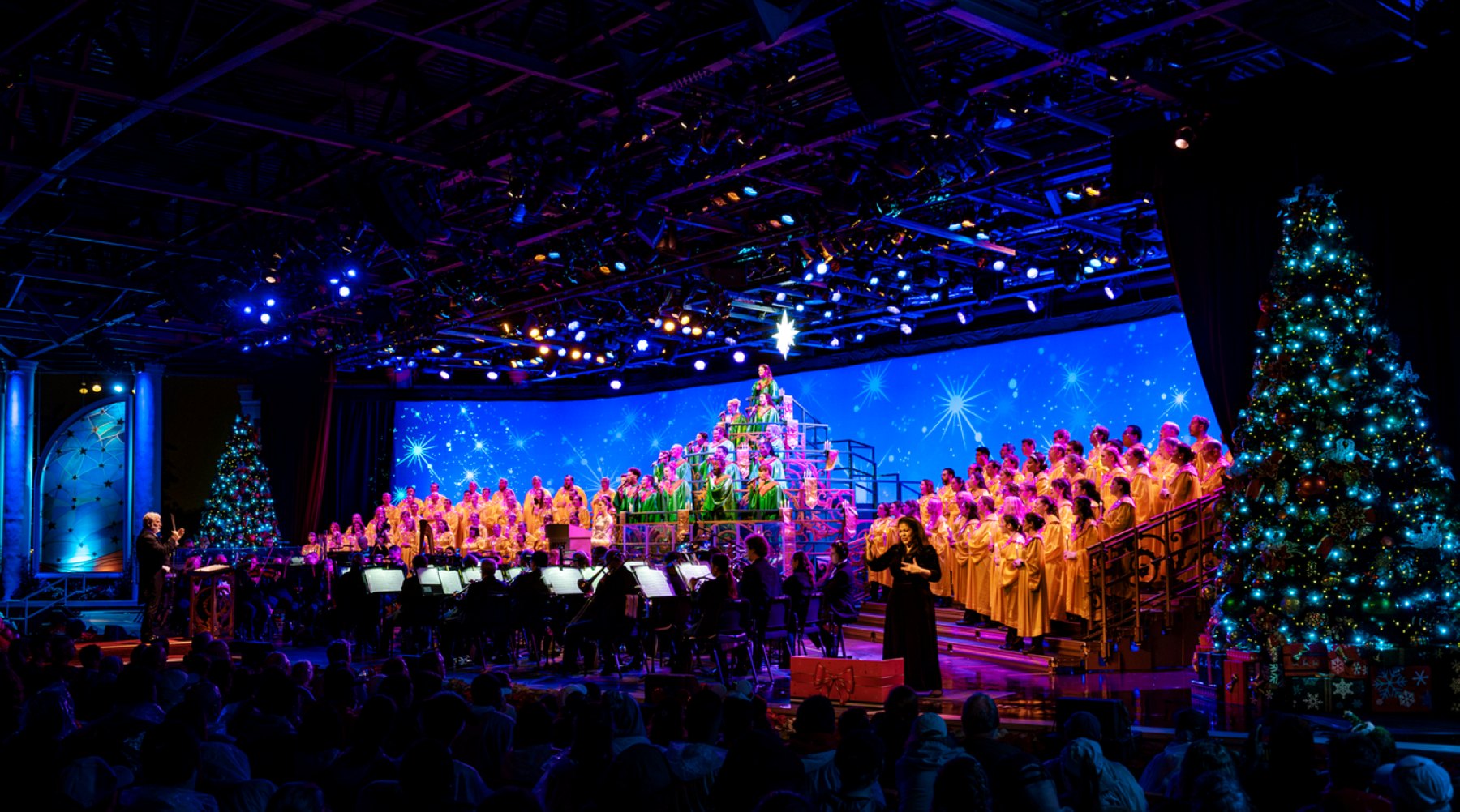 The Candlelight Processional at EPCOT's America Gardens Theatre, with a conductor at left, the orchestra seated across the front of the stage, choir members in green robes on a tree shaped riser at center with choir members in gold robes on the flanking risers, a performer in a black gown standing at the stage front, and lit Christmas trees at either side.
