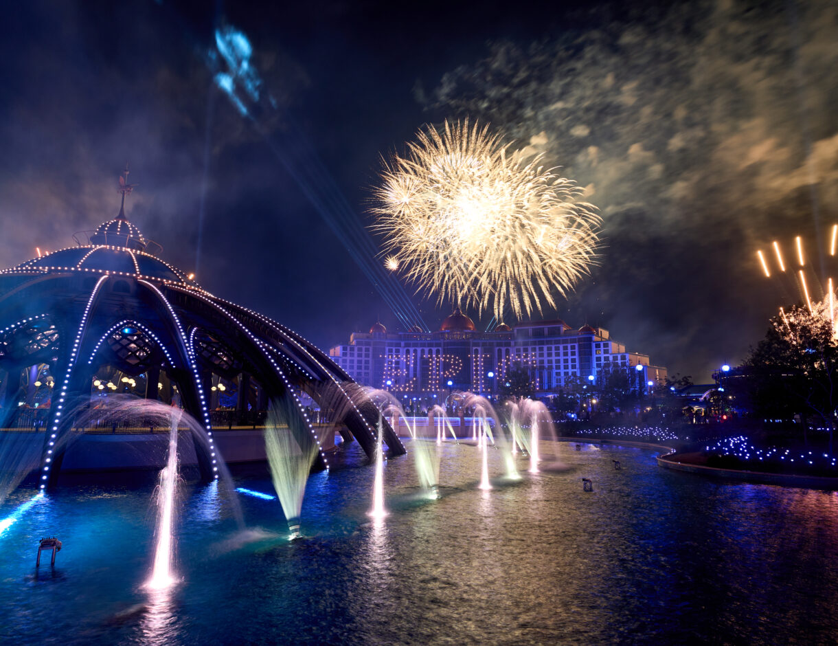 Fireworks burst over the Helios Grand Hotel at night, its windows lit to spell EPIC, with the illuminated fountains of Celestial Park and a domed carousel canopy in the foreground