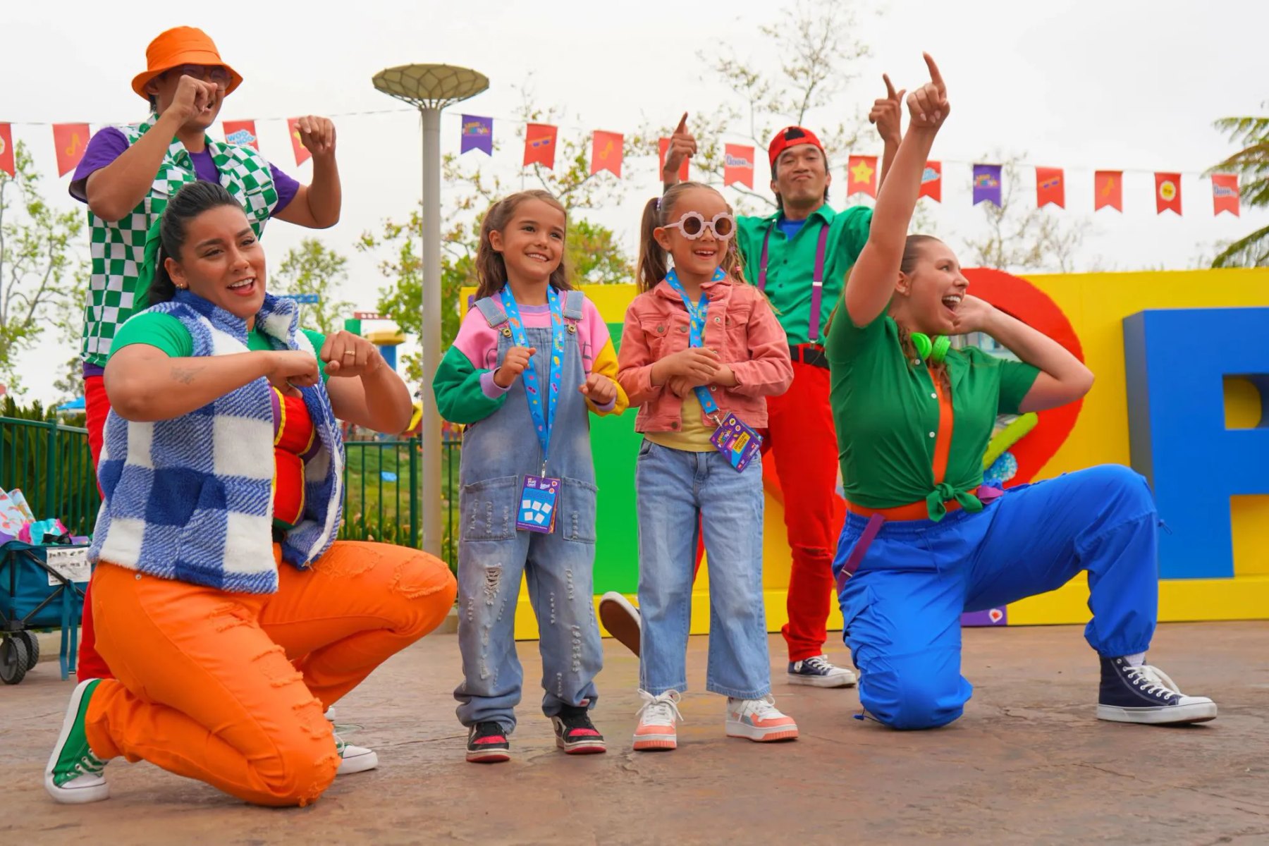 Four costumed Legoland performers in bright green, orange, red and blue outfits posing mid dance with two young girls wearing lanyards, in front of large colorful three dimensional letters on a yellow wall, with printed pennant flags strung overhead.