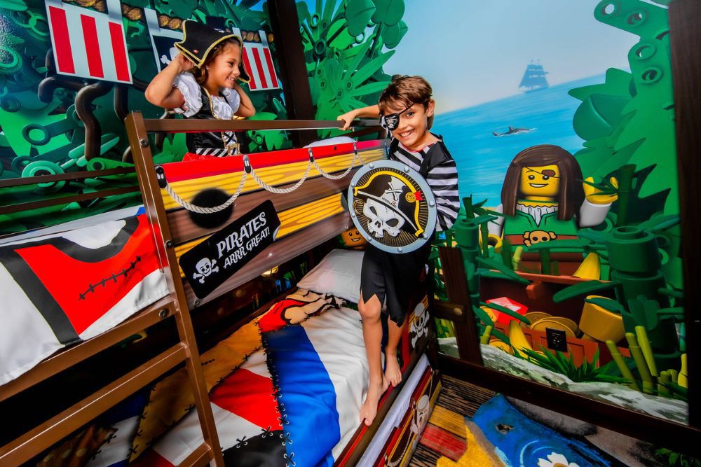 Two children in pirate costumes on wooden bunk beds in a Pirate Island Hotel room, a girl in a tricorn hat on the top bunk and a boy in an eye patch holding a skull shield below, against a painted LEGO jungle mural with a giant pirate minifigure, a tall ship and a shark