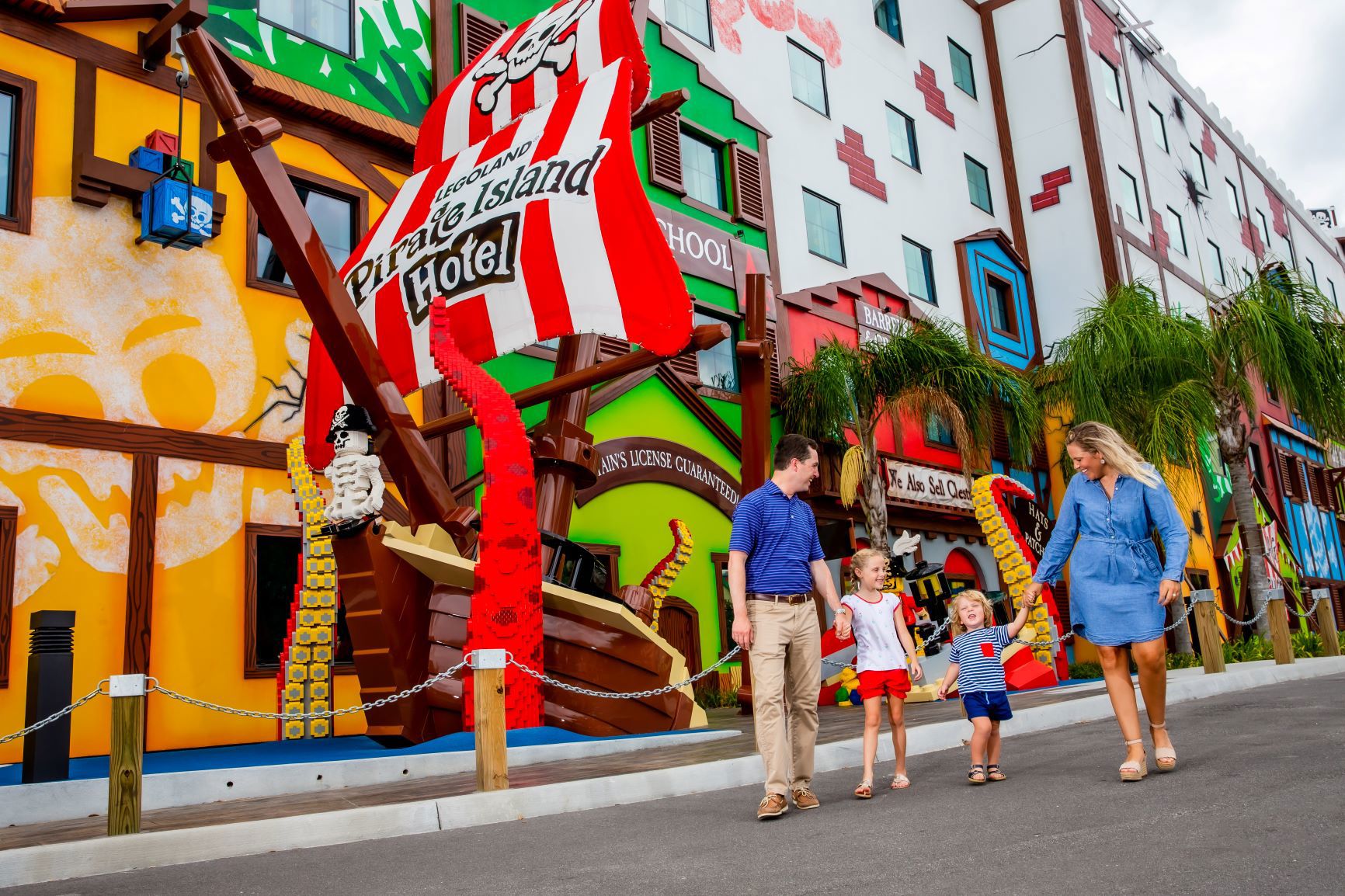 A family of four walks along the road outside Pirate Island Hotel past a large LEGO pirate ship facade whose red and white striped sail reads LEGOLAND Pirate Island Hotel, with a LEGO skeleton pirate on the rigging and palm trees along the colourful hotel block behind