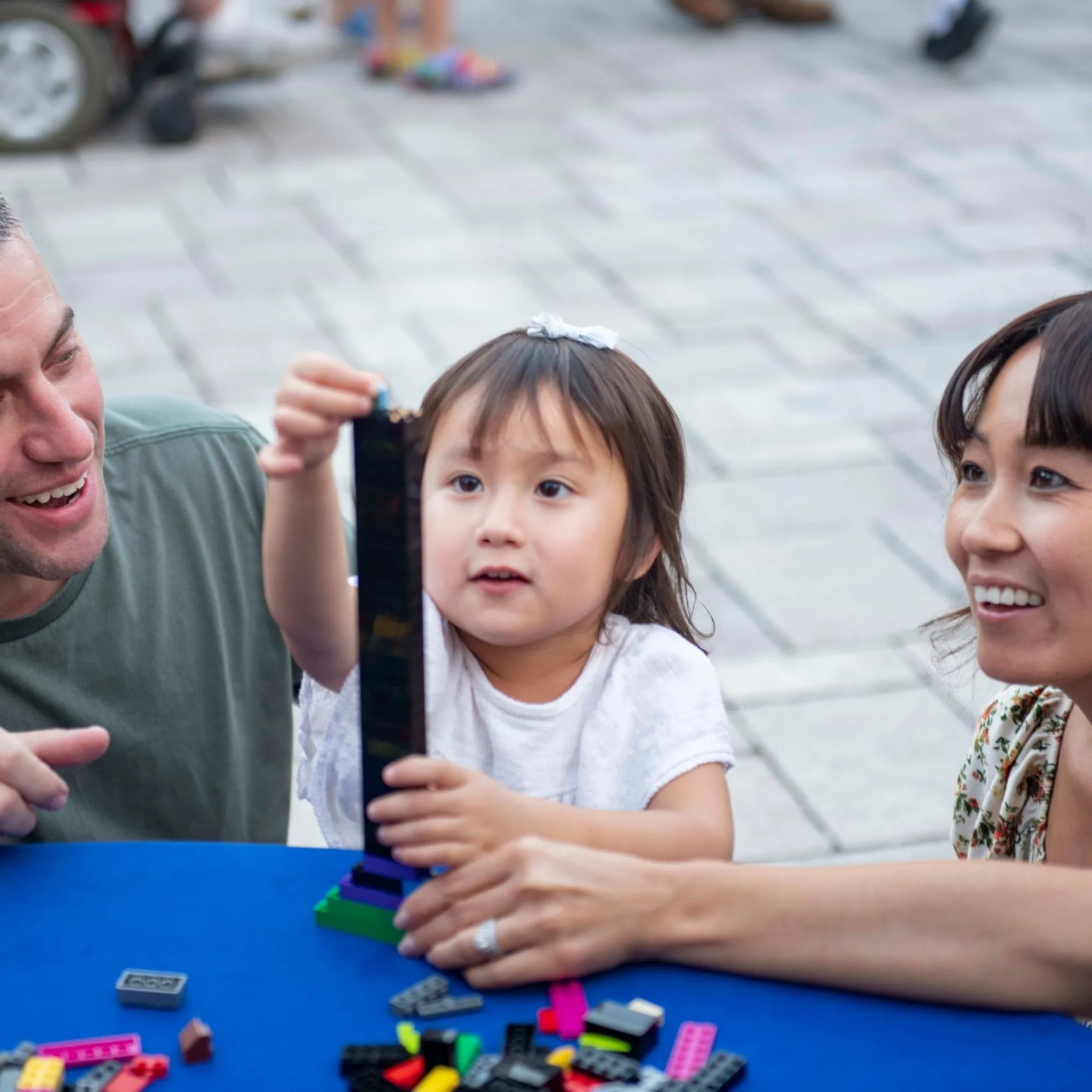 A young girl in a white shirt standing at a blue build table, holding up a tall tower of black LEGO bricks on a green and purple base, with a smiling man at the left of the frame and a smiling woman at the right, and loose colored bricks scattered across the table in front of her.
