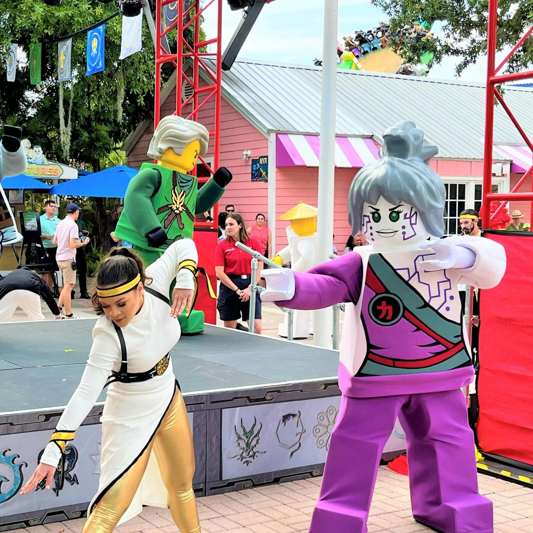 An outdoor stage show at Legoland Florida, with a performer in a white and gold ninja costume bending forward at left, a costumed LEGO character in green behind her, a costumed LEGO character in purple and white with silver hair at right, a staff member in a red polo shirt watching, and a roller coaster train passing above the rooflines behind.