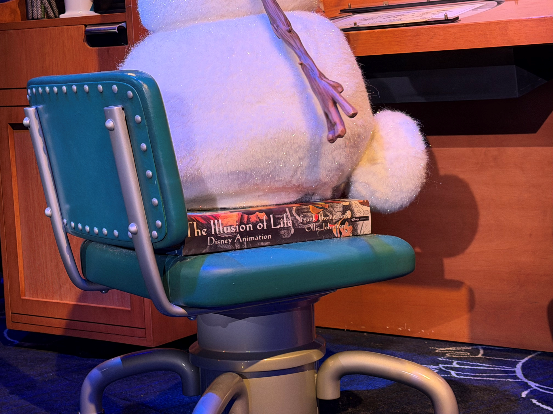 Close-up of the lower half of an Audio-Animatronics Olaf figure perched on a hardback copy of The Illusion of Life by Frank Thomas and Ollie Johnston, which rests on a teal padded animator's chair in front of a wooden animator's desk