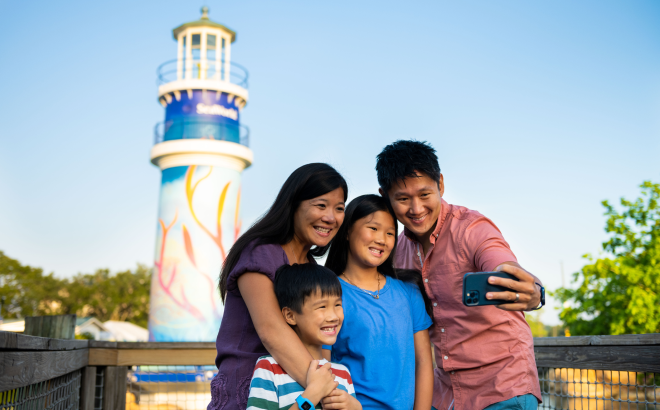 A family of four poses for a phone selfie on a wooden walkway with the painted SeaWorld lighthouse rising behind them under a clear sky