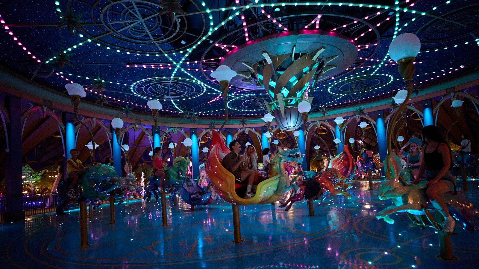 The Constellation Carousel in Celestial Park at night, its creature mounts lit from within, riders aboard, beneath a domed canopy strung with green and pink lights.