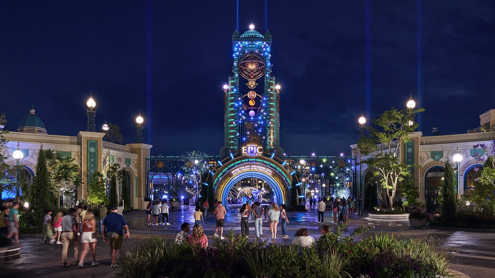 The illuminated Epic Universe entrance portal at night, with the Universal Epic Universe sign on the central tower, searchlight beams overhead and guests crossing the plaza toward the archway.