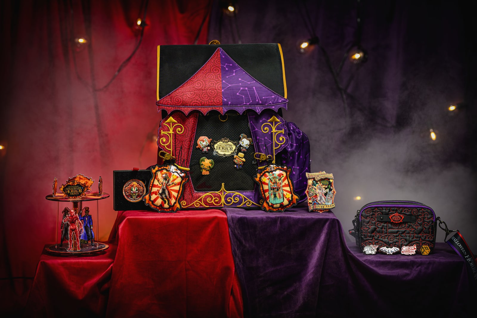 A studio display of Halloween Horror Nights merchandise on red and purple velvet: a circus tent shaped backpack with character pins on its mesh front, ornate carnival frame plaques, a quilted crossbody bag with enamel pins lined up in front of it, and a lit two-tier carousel display holding character figures.