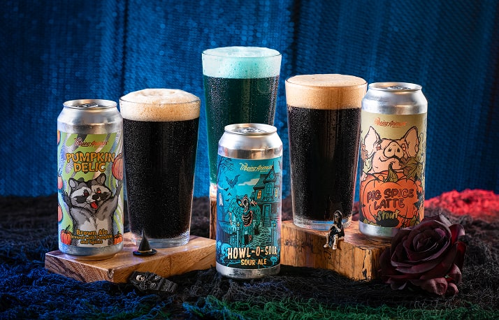 Three Tripping Animals beer cans and three filled glasses arranged on and around wooden blocks against dark blue fabric: Pumpkin Delic with raccoon artwork, Howl-O-Sour with haunted house artwork in front of a bright teal poured glass, and Pig Spice Latte Stout with a pig behind a pumpkin. The other two glasses hold dark beer.