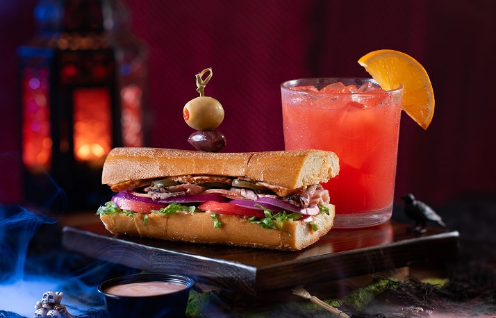 A loaded hoagie sandwich on a wooden board with an olive-topped cocktail pick, a red iced drink garnished with an orange wedge, a small black crow figurine and a cup of pink aioli, a glowing red lantern behind and blue-lit fog at the front left.