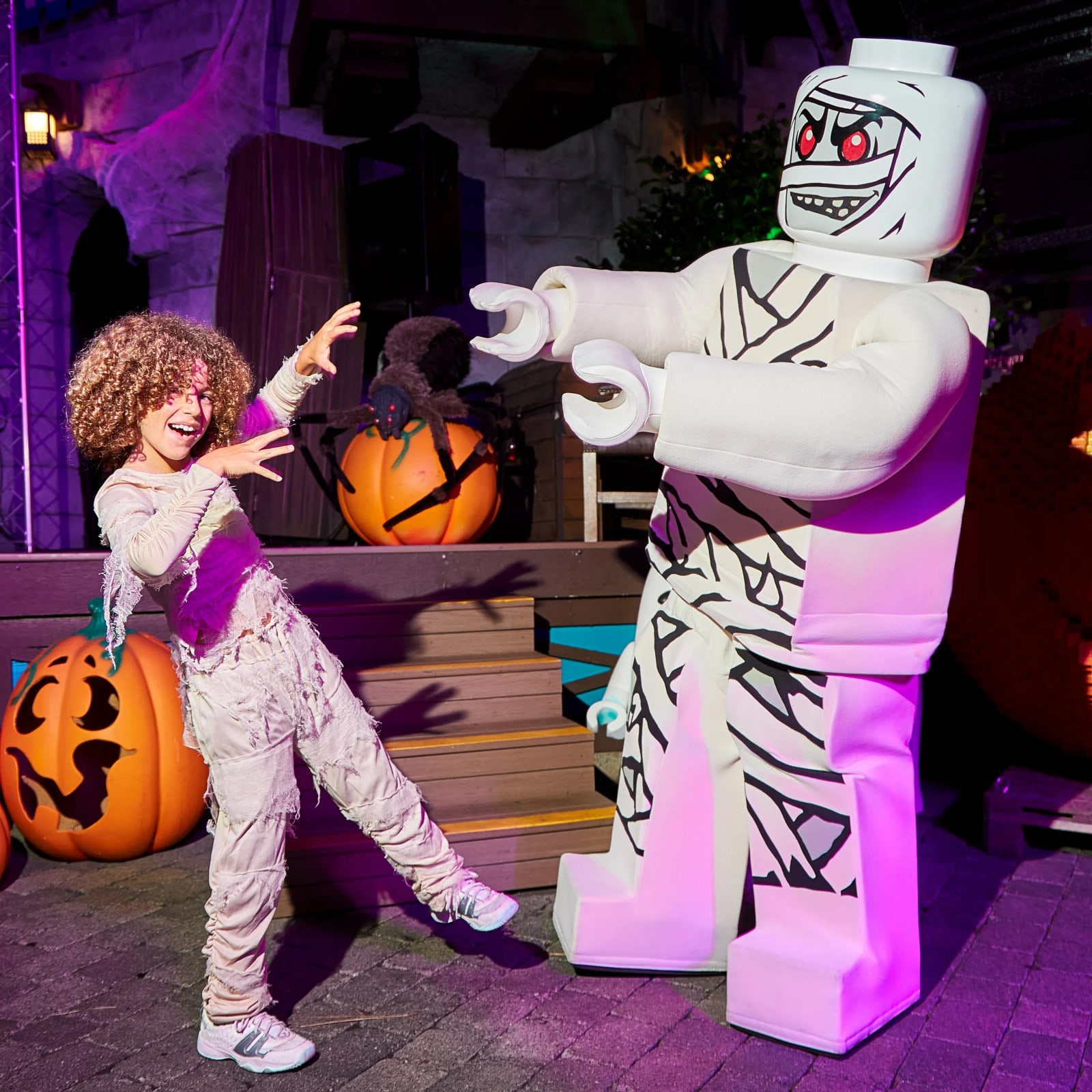 A child in a tattered mummy costume playfully raises their hands toward a tall walkaround LEGO Mummy character with red eyes and printed bandages, at night, with carved pumpkins and a spider prop nearby.