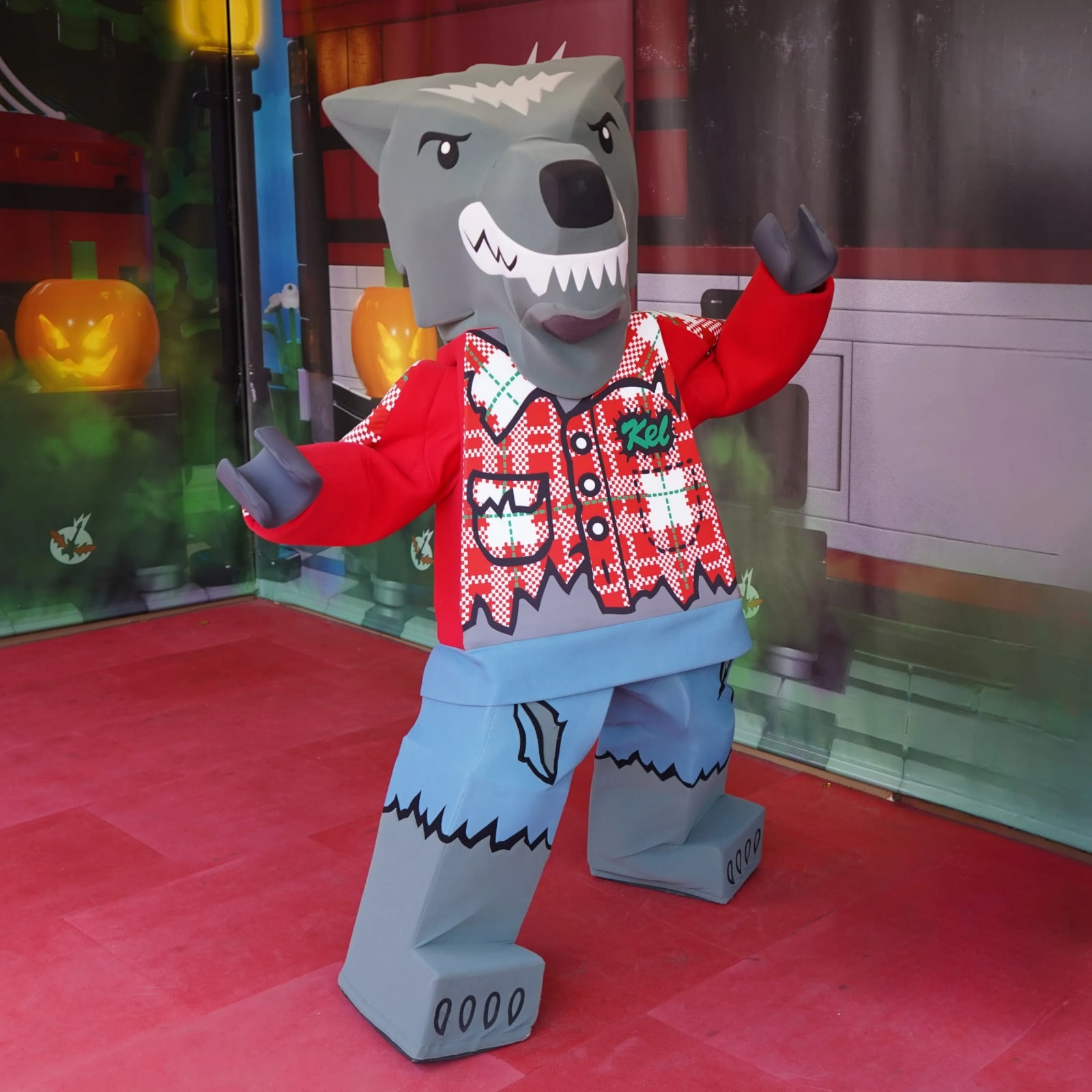 The walkaround LEGO Wolf Guy character standing on brightly lit red pavement, with a grey wolf head, bared teeth, a red plaid shirt with a green patch reading Kel, and ripped blue jeans.
