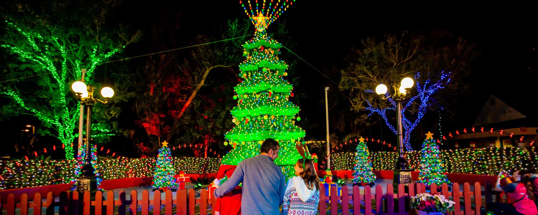 At night, a man in a grey sweater and a girl wearing a reindeer antler headband stand with their backs to the camera behind a red picket fence, facing a towering LEGO brick Christmas tree covered in green lights, coloured baubles and a gold star, with trees wrapped in green and blue lights on either side and a field of multicoloured lights behind them.