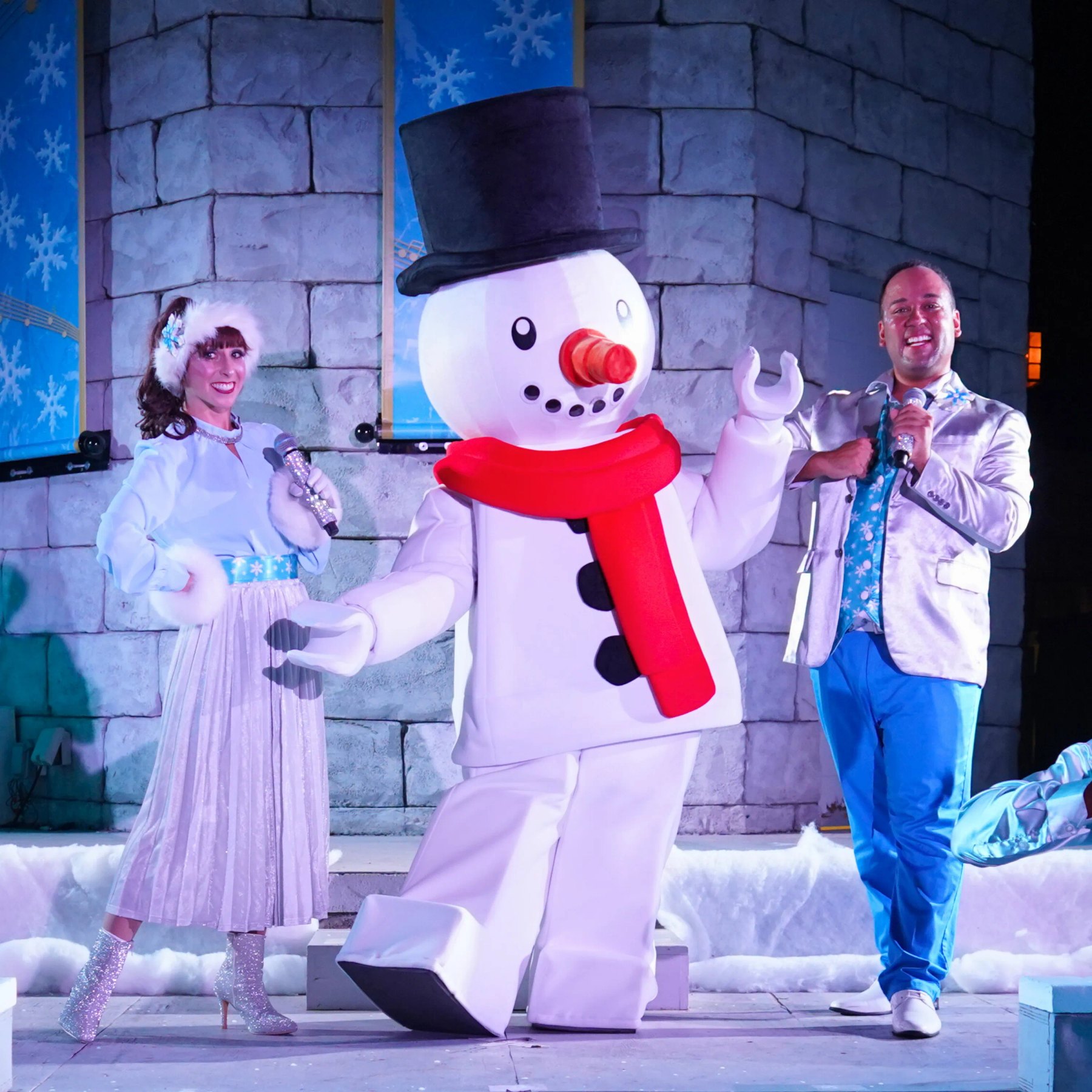 A performer in a white and silver outfit with a snowflake hair piece holds a microphone at left, a walkaround LEGO Snowman with a black top hat, red scarf, black button eyes and an orange carrot nose stands at center with one arm raised, and a performer in a silver jacket and blue trousers holds a microphone at right, on a stage in front of a grey stone wall hung with blue snowflake banners.