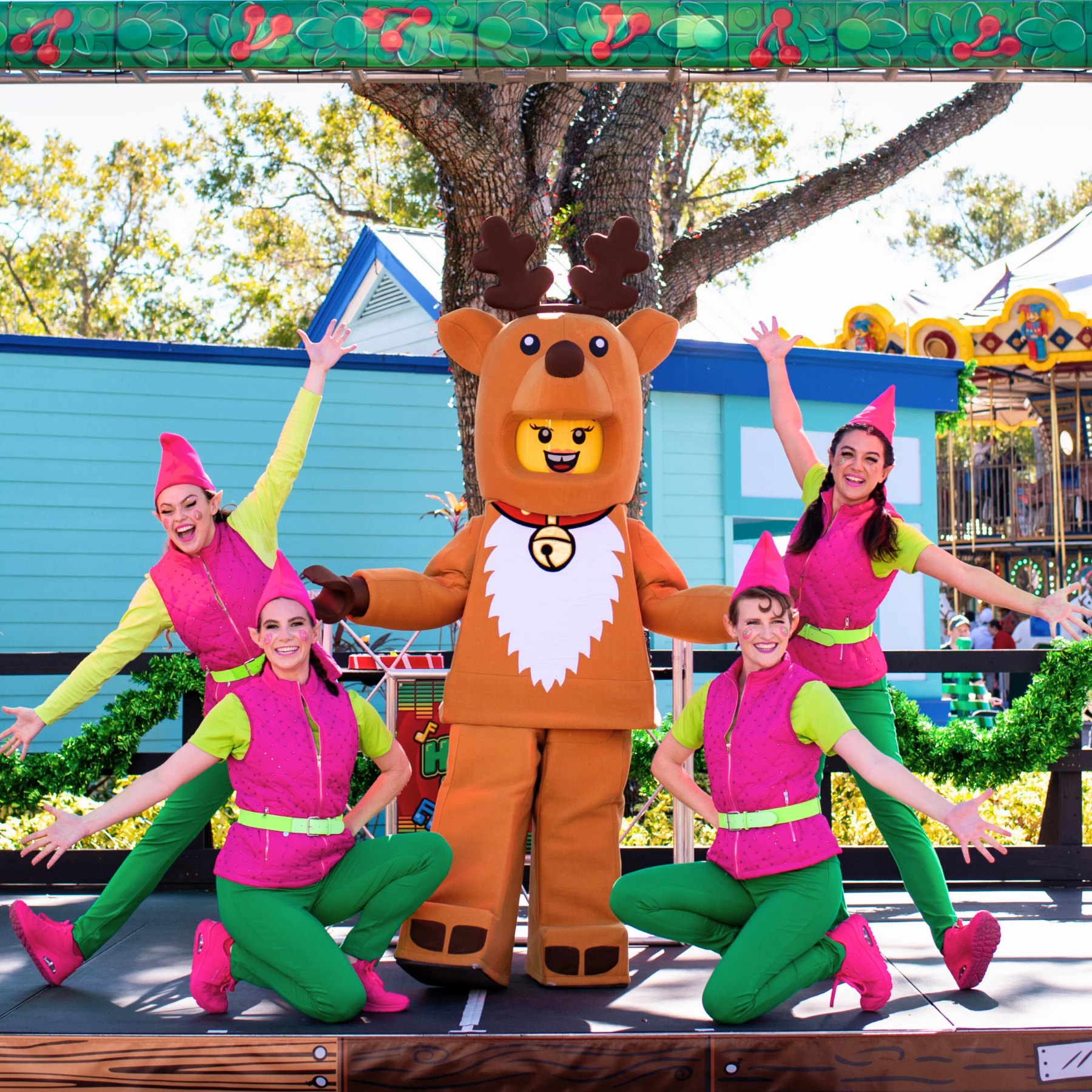 Four performers in hot pink elf hats and vests with lime green sleeves and green trousers strike a pose on an outdoor stage, two kneeling and two standing with arms flung wide, around a walkaround LEGO Reindeer Girl character in a brown reindeer costume with antlers, a white chest ruff and a gold bell collar, with a carousel visible behind them.