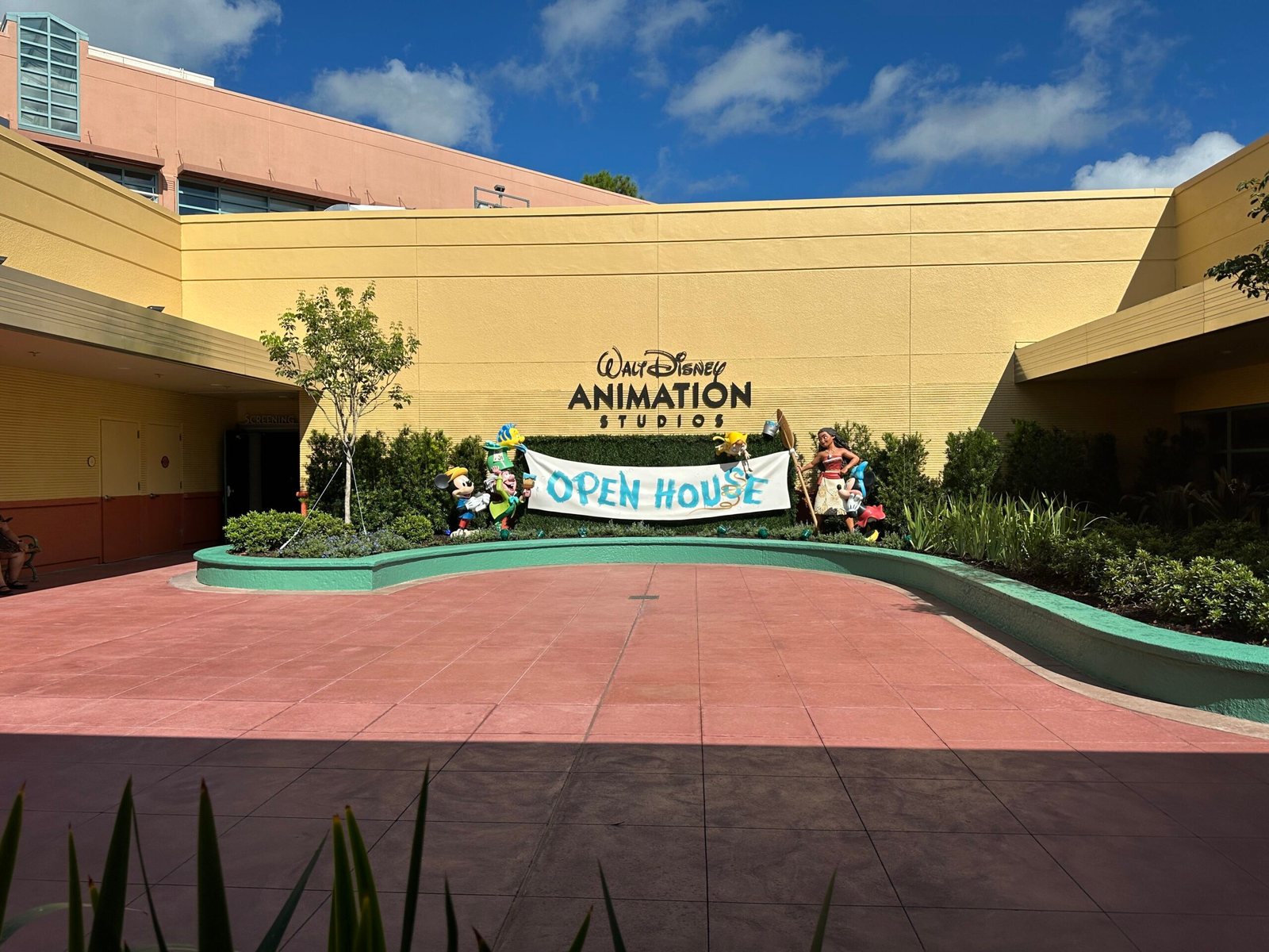 The exterior courtyard of the Walt Disney Animation Studios building at Hollywood Studios, with a painted OPEN HOUSE banner strung between figures of Mickey Mouse, the Mad Hatter with Flounder perched on his hat, Pinocchio, Moana and an upside down Minnie Mouse, below the studio wordmark.