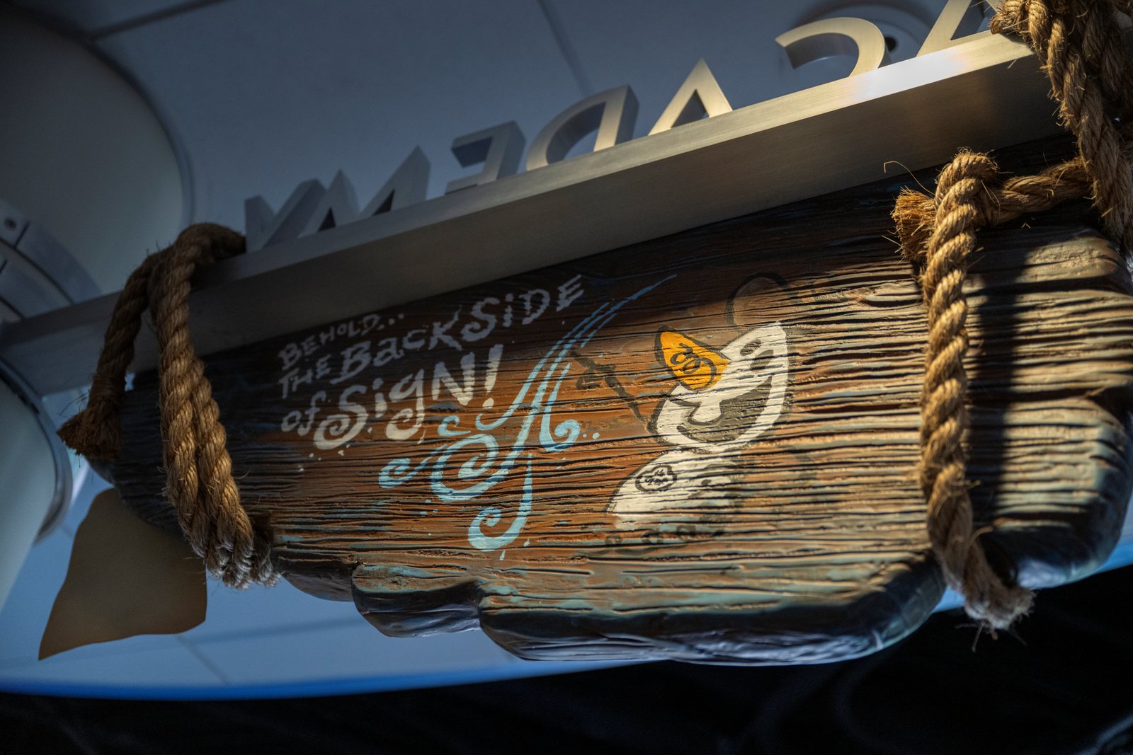 The back of the hanging wooden Olaf Draws! marquee, hand painted with the words BEHOLD... THE BACKSIDE of SIGN! beside a doodle of Olaf's face, with rope ties at each end and the reversed ACADEMY letters on the wall behind it.