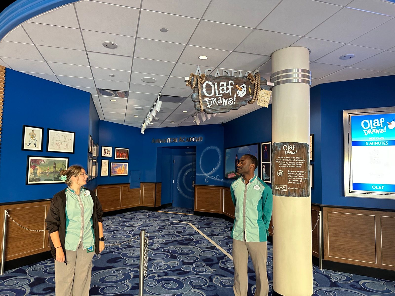 The Olaf Draws! entrance area, with a hanging wooden sign, a wall of framed animation art, an information plaque and a digital board reading NEXT CLASS 5 MINUTES, with two cast members standing at an empty chained queue.