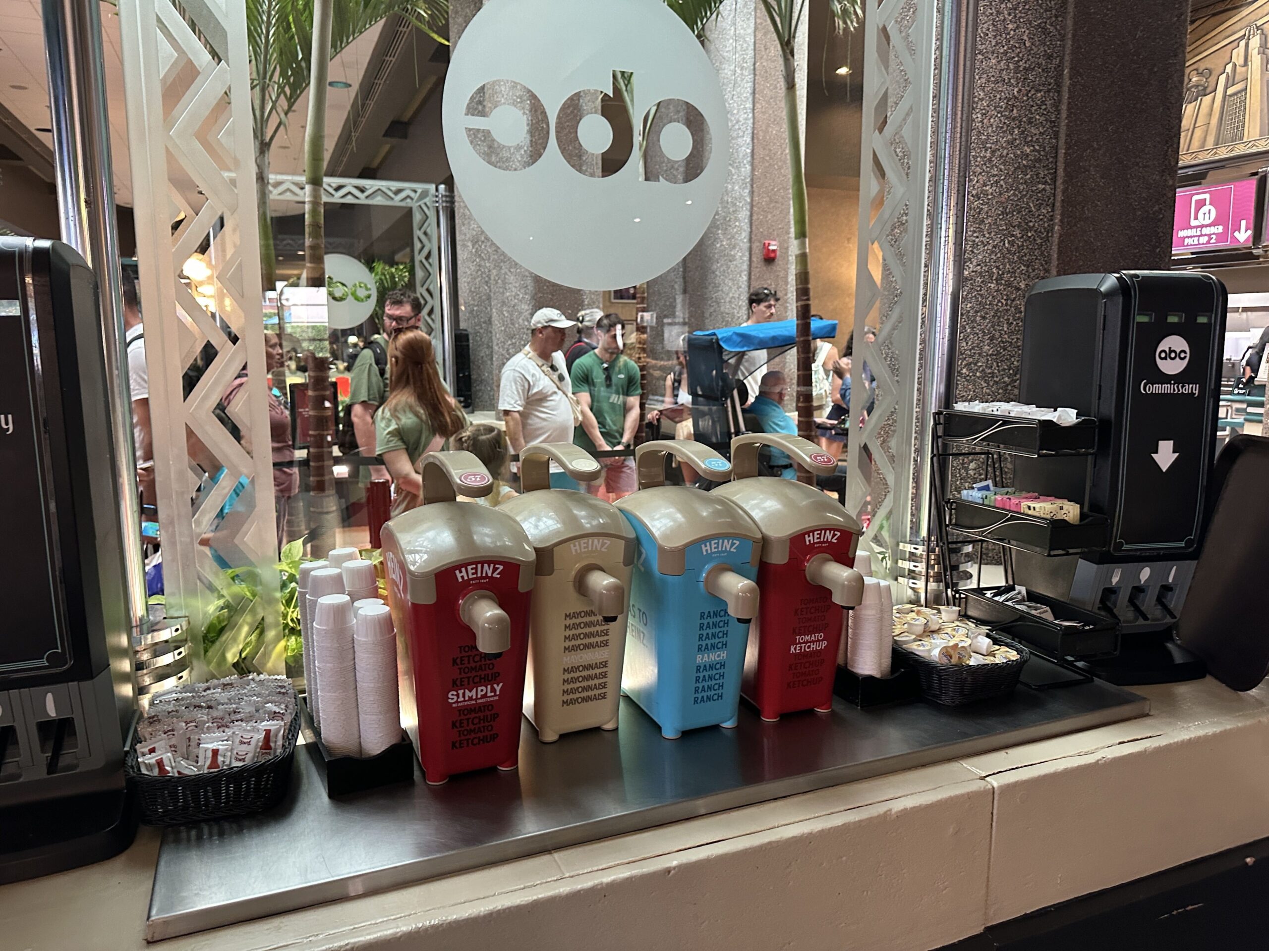Four Heinz pump dispensers in a row on a stainless steel counter at a theme park quick service restaurant, in front of a glass partition. From left, a red Simply Tomato Ketchup unit, a tan Mayonnaise unit, a blue Ranch unit and a red Tomato Ketchup unit, all with tan lids and spouts. Stacks of white portion cups sit beside them, a basket of hot sauce packets is at far left, and a black sign reading abc Commissary with a down arrow and a pink Mobile Order Pick Up 2 sign are at right, with the frosted abc logo roundel and guests visible behind.