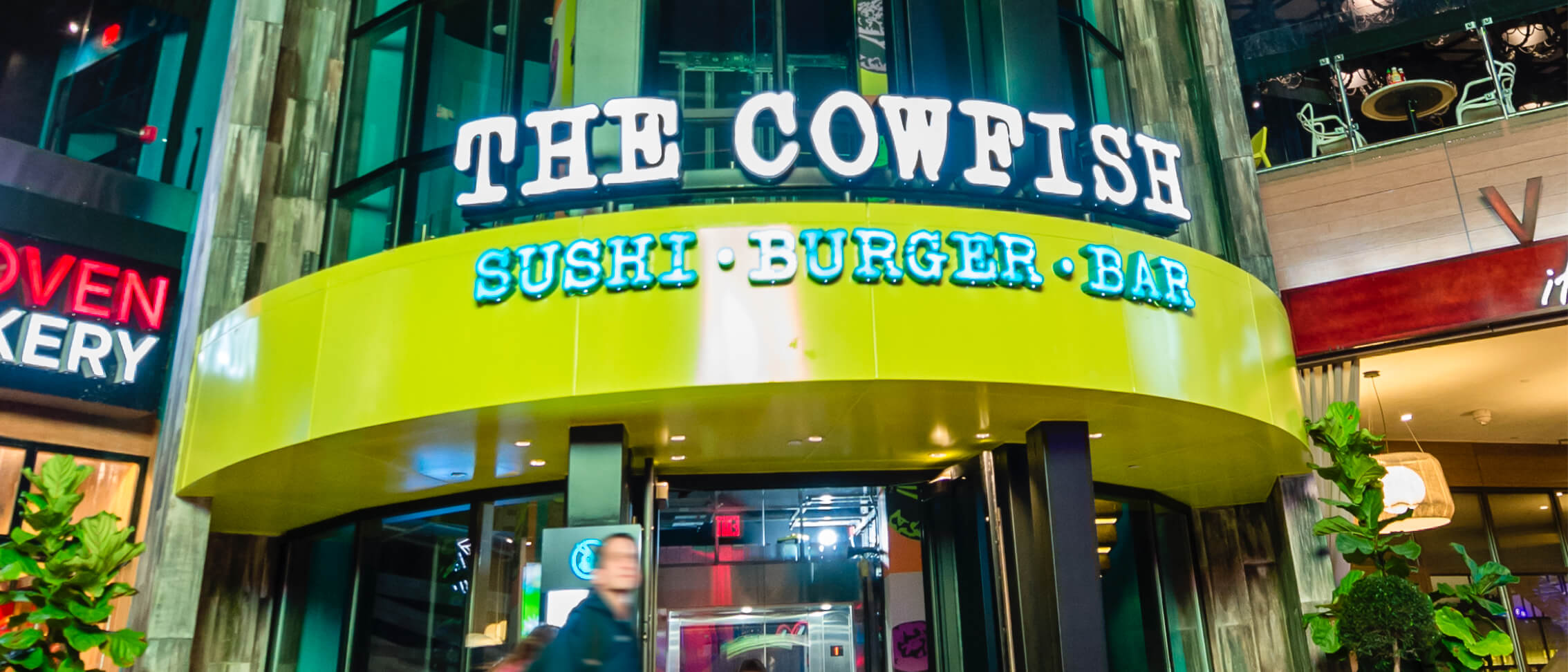 Night view looking up at the curved entrance rotunda of The Cowfish at Universal CityWalk. The restaurant name is in large white block capitals above a glossy lime green band reading Sushi, Burger, Bar in pale blue outlined letters. A guest in a dark jacket walks past the glass doors below, with a red neon bakery sign at the far left.