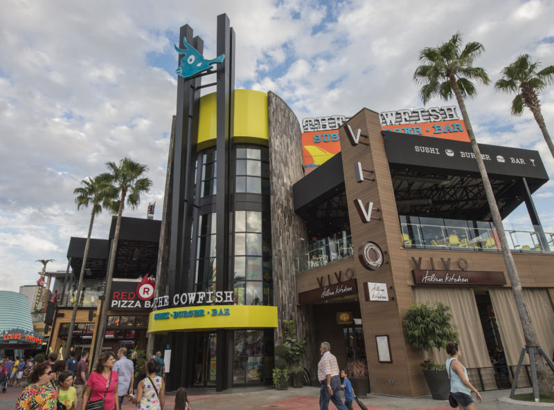 Daytime wide view of the Universal CityWalk block, with a blue cow-fish sculpture mounted on a black tower above a yellow drum and signage reading The Cowfish Sushi Burger Bar. VIVO Italian Kitchen's wooden facade with a tall vertical VIVO sign stands at the right, a pizza bakery sign at the left, and families in summer clothes walk the paved promenade under palms and a partly cloudy sky.