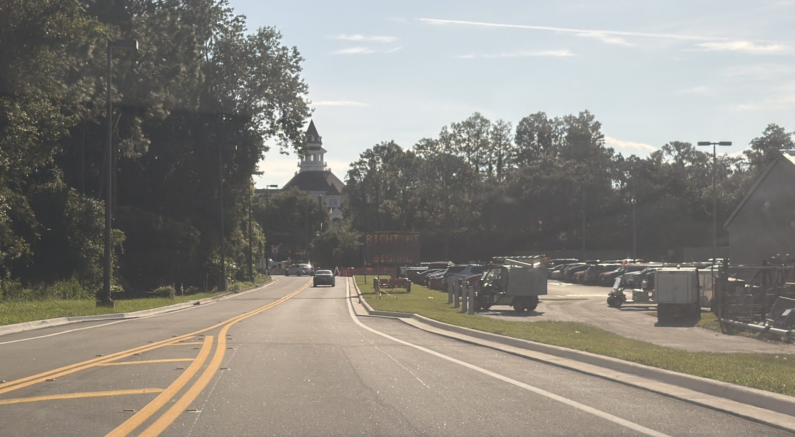 A two lane road with double yellow center line, an amber LED board reading RIGHT AT SIGNAL beside a parking lot full of cars and equipment, and a Victorian rooftop with a cupola in the distance