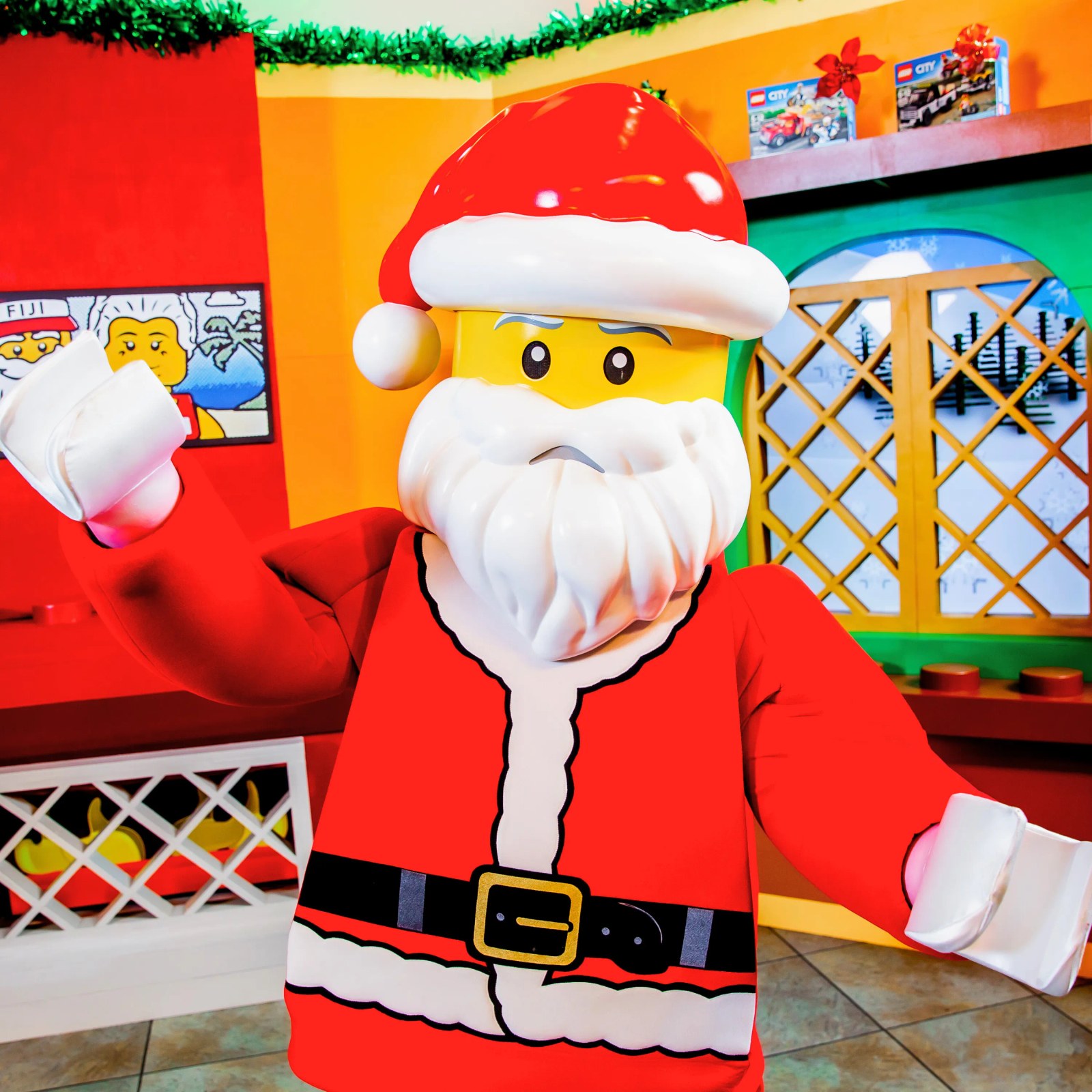 A LEGO Santa walkaround character with a molded yellow minifigure face and white beard waving inside a brightly colored room with LEGO City boxes on a shelf