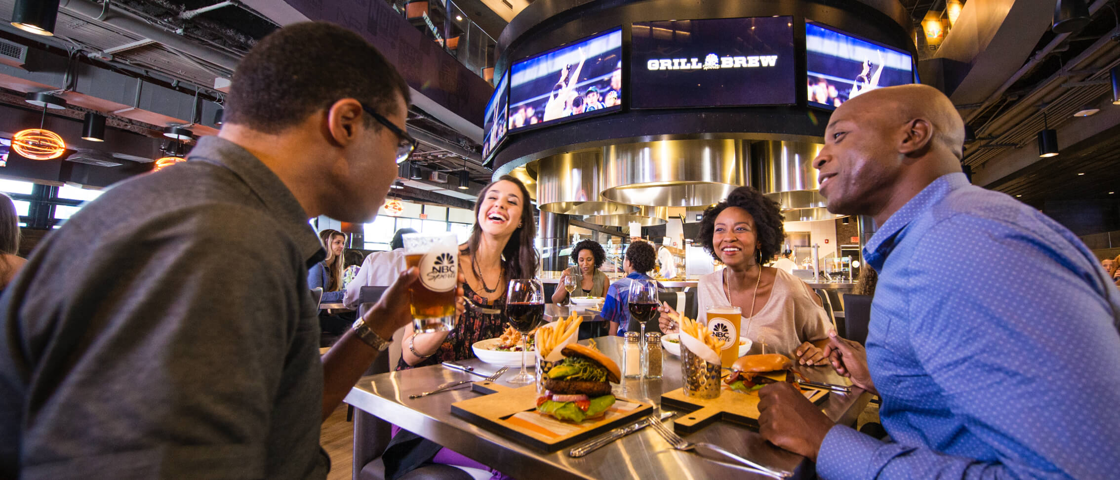 Interior of NBC Sports Grill and Brew. Four adults sit around a steel topped table mid conversation, one holding an NBC Sports branded pint glass and a second such glass standing on the table, with two glasses of red wine, burgers served on pale wooden boards with dark edges and fries in dimpled metal cups in front of them. Behind them a circular overhead video ring reads Grill and Brew above a lit open kitchen.