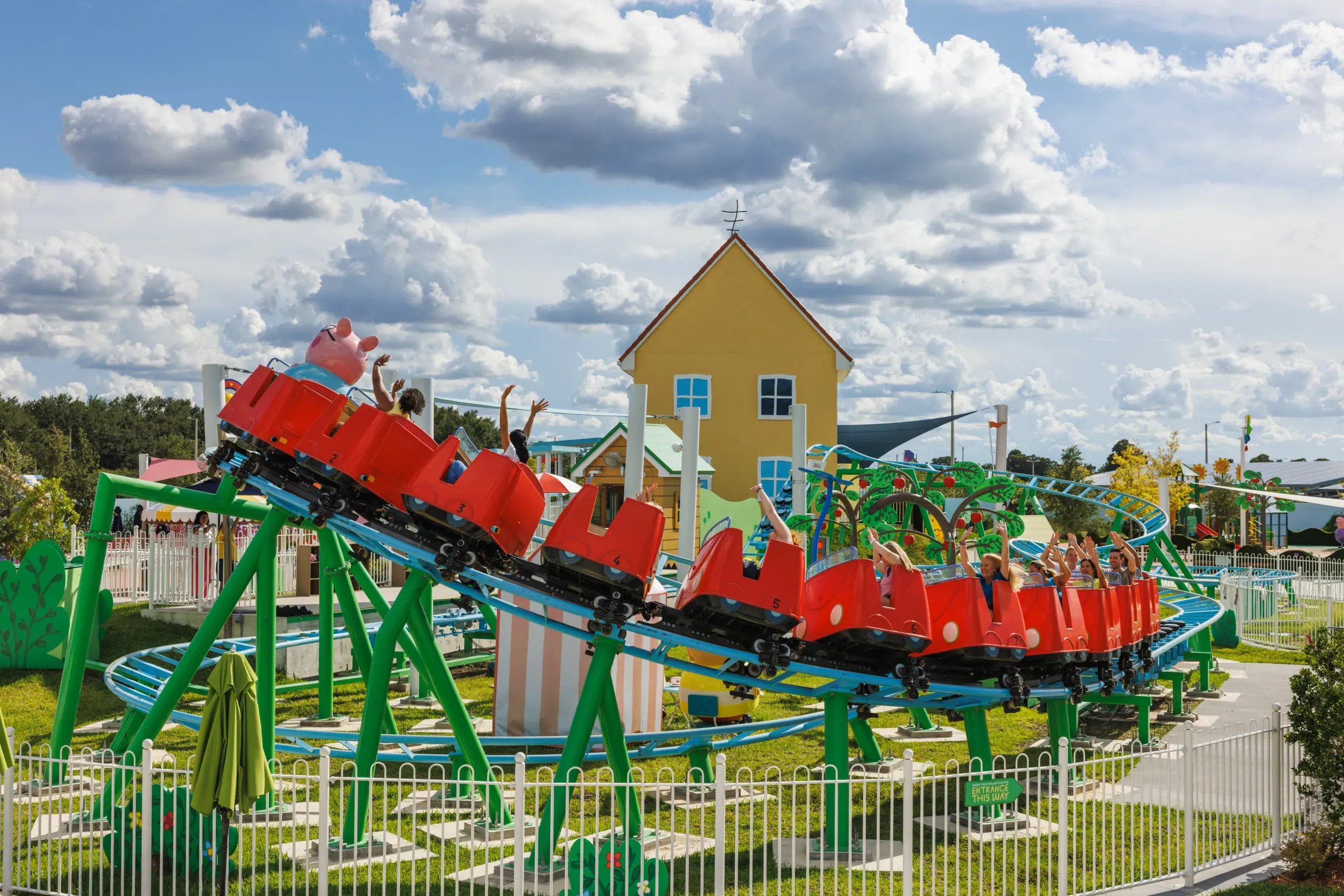 Daddy Pig's Roller Coaster running at Peppa Pig Theme Park, a red train of numbered cars full of riders with their hands up, on blue track with green supports, with the yellow Peppa house behind it and a white picket fence in front