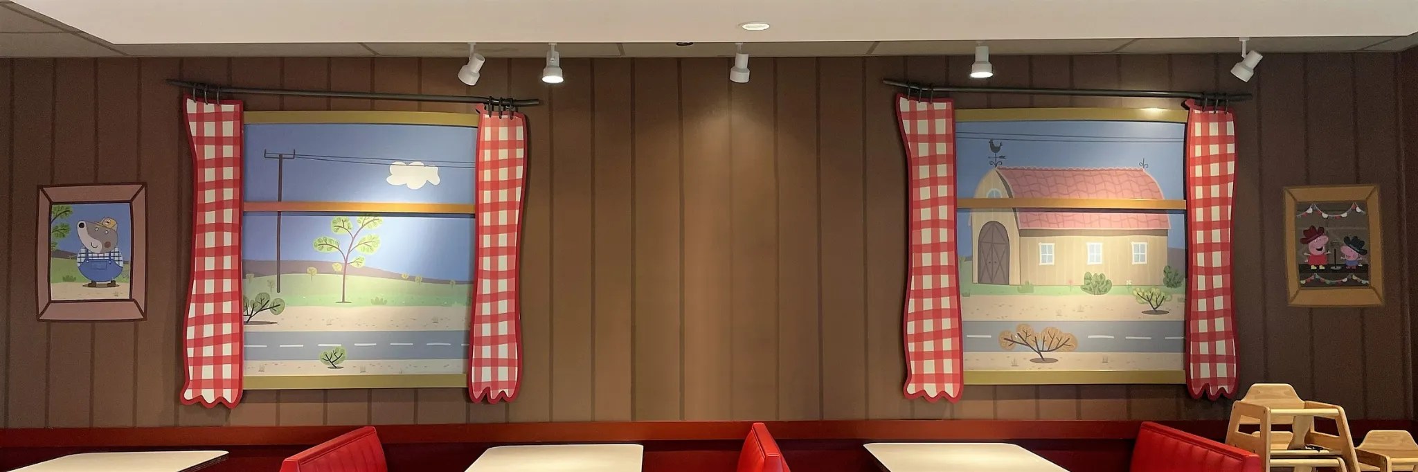 Interior of a diner at Peppa Pig Theme Park with brown wood panel walls, two painted countryside windows framed by red gingham curtains, red booth seating with pale tabletops and a wooden high chair at right